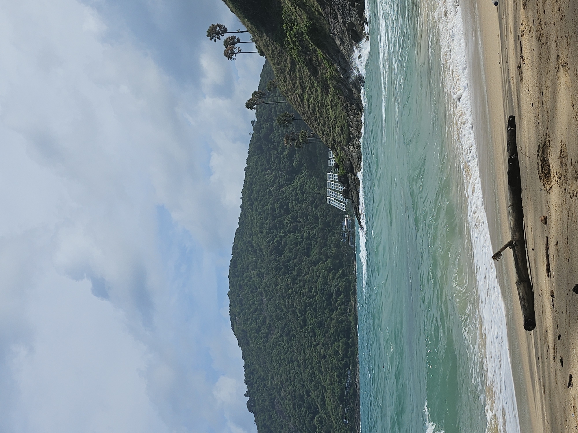 The image depicts a scenic view of a beach in Rawai, Thailand, under a cloudy sky. In the foreground, a sandy beach is visible with a weathered log lying horizontally across it. The ocean stretches out to the horizon, with turquoise-colored water and gentle waves breaking on the shore. Along the coastline, a lush green hill rises steeply, dotted with palm trees and vegetation. In the middle ground, a white building complex with a distinct architectural style is nestled against the hillside. Rocks and rough terrain are present along the water's edge, with some waves crashing against them. The overall scene is one of natural beauty and coastal serenity, with a touch of human development.