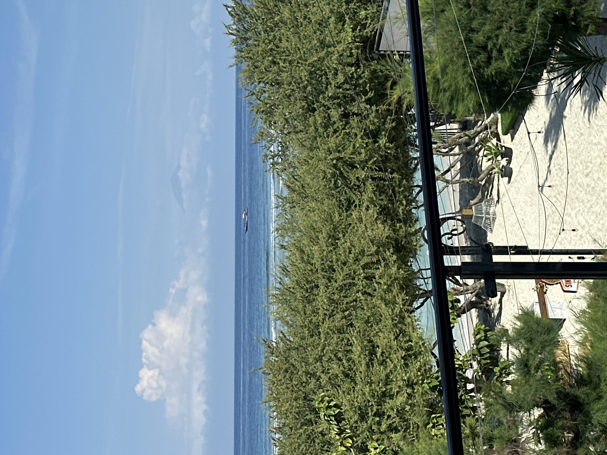The image presents a view of the sea and a beach in an unidentified location, likely in Indonesia, as indicated by the city name. The sea is visible in the distance, merging into the sky under a blue sky with some clouds. Closer to the foreground, dense green trees obscure a significant portion of the scene. Through the trees, glimpses of a sandy beach can be observed, dotted with beach furniture such as loungers and chairs. Above the beach area, there are strings of lights. The foreground also includes a metal frame, possibly part of a structure or equipment, which adds a man-made element to the natural scene.