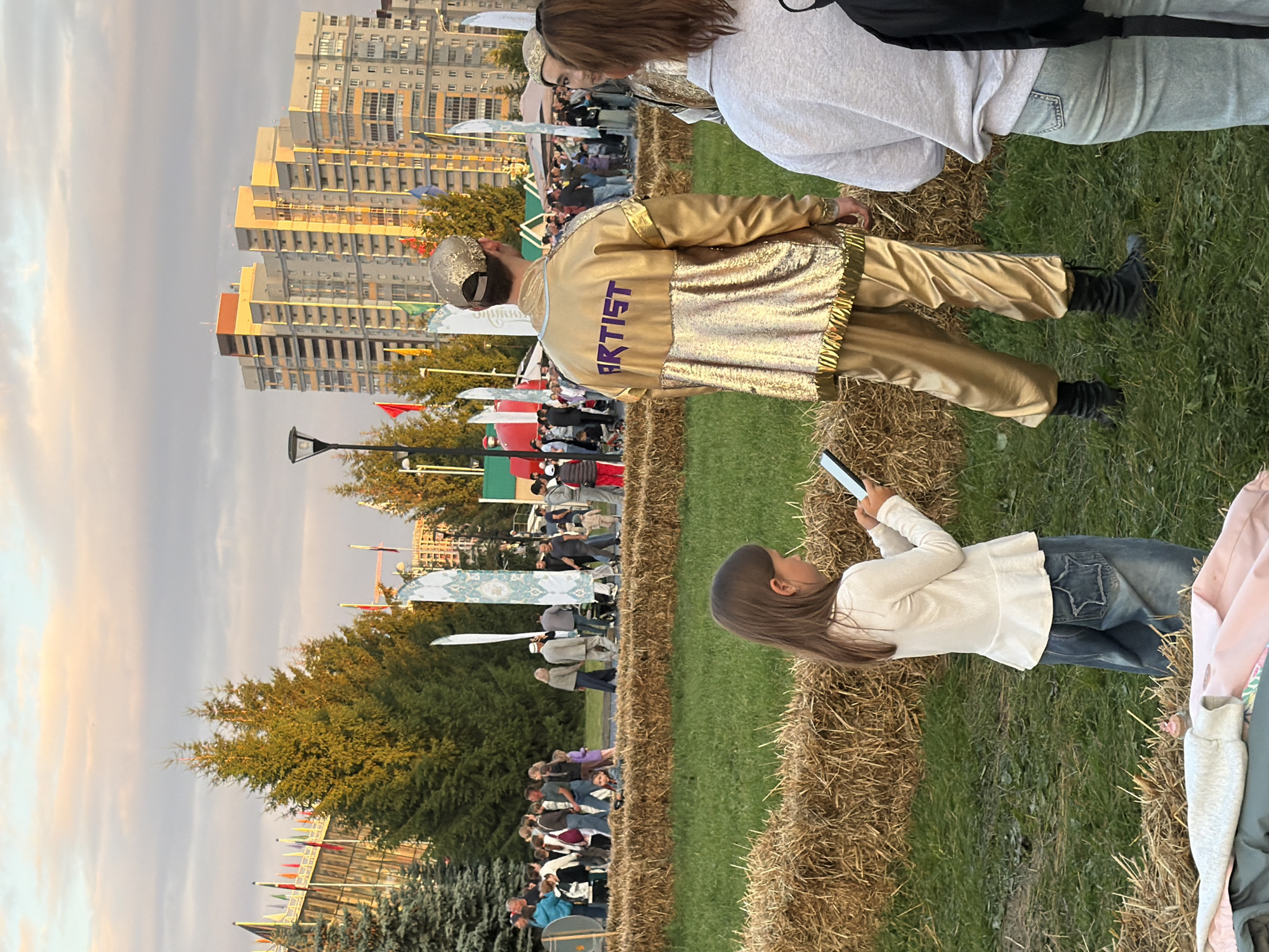 In an outdoor urban setting, likely a park or festival, several people are gathered. In the foreground, a young girl with long brown hair is looking down at a tablet she is holding. She is wearing a white long-sleeved shirt and blue jeans with a star-shaped design on the back pocket. To her right and slightly behind her, a person in a shiny gold tracksuit with "ARTIST" written in purple on the back is facing away from the camera, looking towards a crowd. The tracksuit consists of a jacket and pants, and they are wearing a gold cap and black boots.
Behind these individuals, a diverse crowd is visible, some walking and others standing. They are in front of a line of large hay bales. In the background, there are trees, flags on poles, and tall modern buildings, suggesting an urban environment. The sky above is partly cloudy with some warm-toned light, indicating it might be late afternoon or early evening. The ground is covered with green grass.