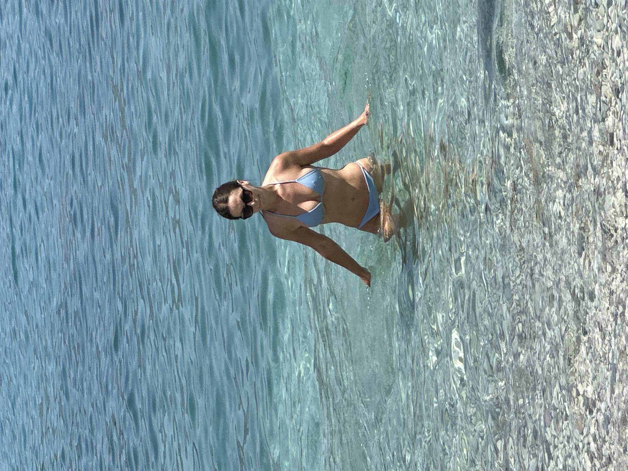 A woman wearing sunglasses and a light blue bikini stands in clear, shallow water. Her hands are resting on the surface of the water, and she appears to be walking or wading. The seabed is visible through the water, covered in small, smooth stones. The water is calm with gentle ripples, reflecting the sunlight. The background consists of more water, with a darker blue hue further out. The scene appears to be a beach or a shallow coastal area. This location could be one of the many beaches in Budva, Montenegro, such as Mogren Beach or Jaz Beach.