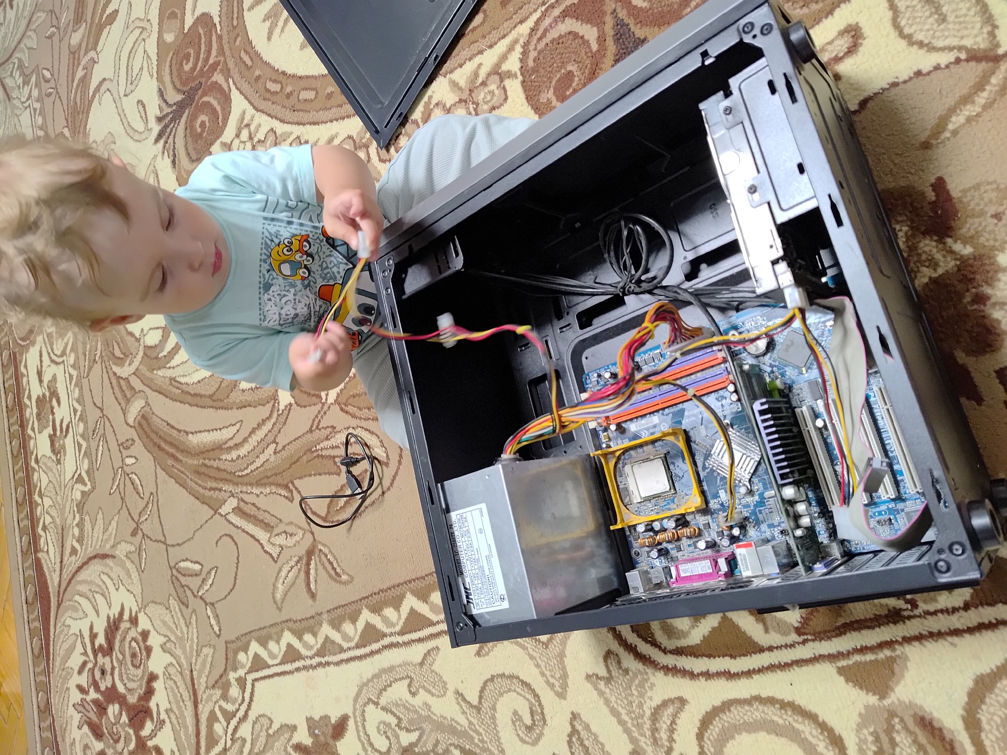 A young child, likely a toddler, is seen from a high angle, sitting on a patterned carpet. The child is wearing a light blue t-shirt with a cartoon design and is interacting with the internal components of an open computer tower. The computer tower is lying on its side on the carpet, revealing the motherboard, power supply, and various cables. The child's hands are manipulating some of the colorful wires. The scene appears to be indoors, given the carpet and the presence of the computer. The lighting suggests daytime. The overall impression is one of a curious child exploring technology. The visible text on the power supply unit reads "MODEL: LC-250ATX P4 INC COMPUTER CO.,LTD. CAUTION!". The location is contextually given as Марковский, Russia.