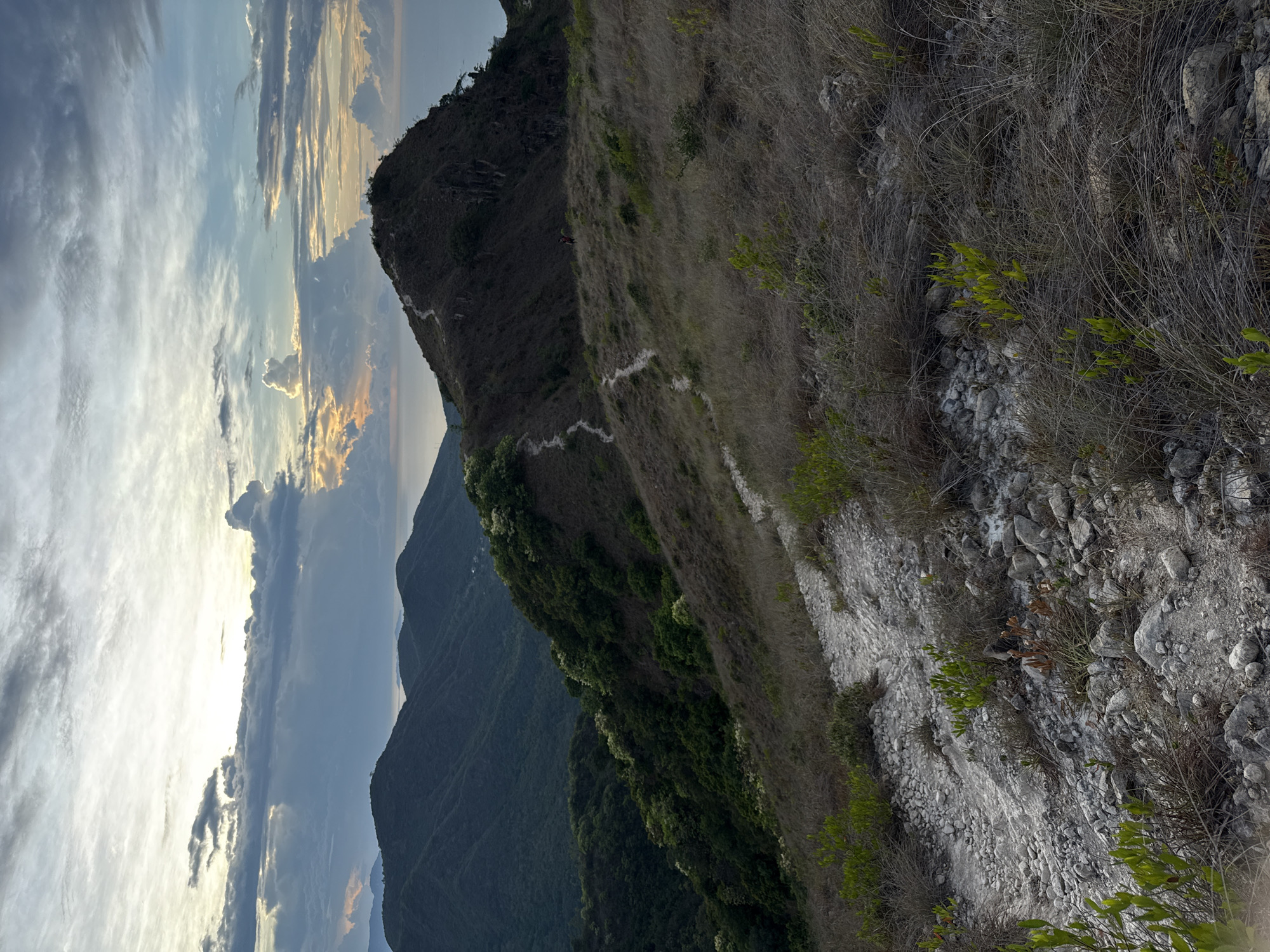 The image depicts a mountain ridge at an angle, with a path winding along the crest. The foreground shows dry vegetation, stones, and small shrubs. The middle ground features a steep mountainside covered in green vegetation. Beyond that are more mountains and a view of the sky with clouds and hints of sunset.
While the description specifies Nha Trang Ward, Vietnam, no elements in the image can verify this location. The presence of the path and vegetation on the ridge are the key defining features. There are no discernible people, objects, actions, or interactions.
