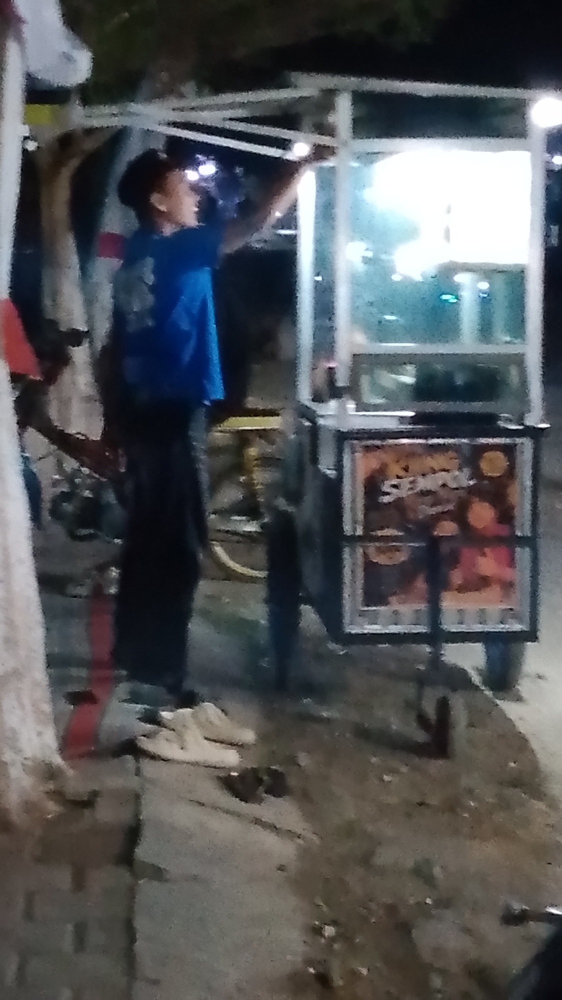 In Karanglangu, Indonesia, at night, a young male wearing a blue t-shirt and dark pants stands near a food cart. He is looking up and appears to be adjusting the cart's lighting. The cart has a glass display case and a sign with text and images, likely advertising the food being sold. In the foreground, on the ground, a pair of light-colored sneakers are visible, along with some debris. The environment suggests an outdoor setting, possibly a street or market area, with artificial lighting illuminating the scene.
