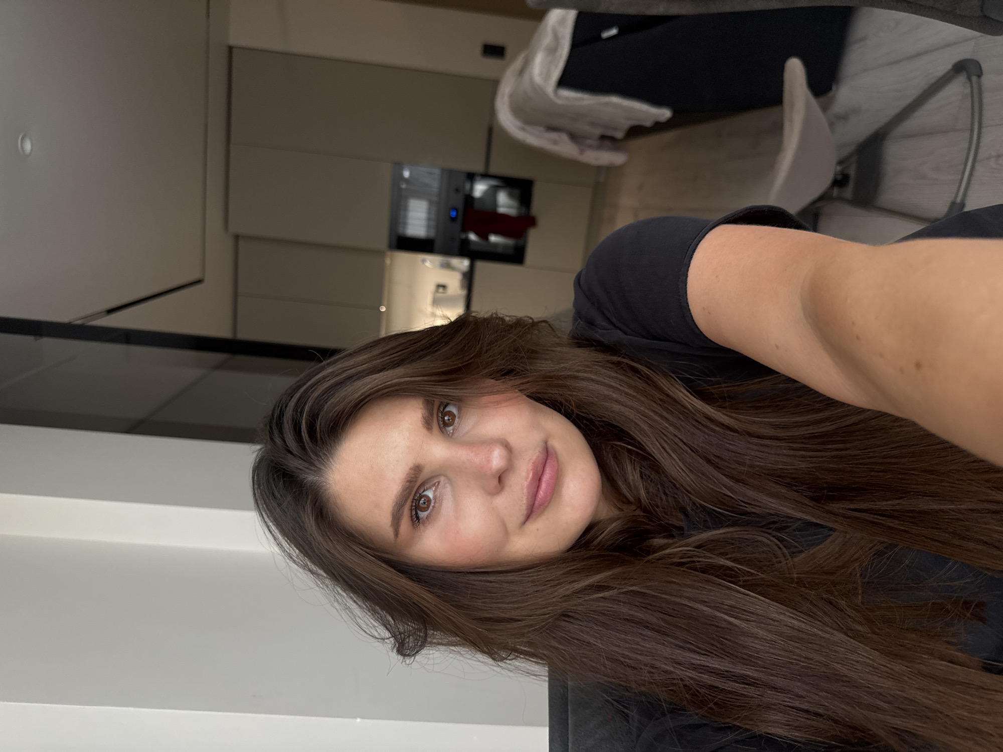 A female individual with long, dark brown hair, fair complexion, brown eyes, and a slight, closed-lip smile takes a selfie, looking directly at the camera. She wears a dark, short-sleeved top, with her right arm prominent in the foreground. The background depicts a modern indoor apartment setting, specifically a combined kitchen and living area in Sofiivska Borshchahivka, Ukraine.
The kitchen features light-toned, flat-panel cabinetry and an integrated dark-front oven with a visible blue display and a red cloth hanging from its handle. A white appliance and a spotlight are visible on the counter. To the right, a dark piece of furniture is partially visible, draped with a light-colored blanket. A light-toned, minimalist-style chair with a curved base sits on light wooden flooring. The ceiling includes recessed lighting, and a dark overhead panel defines part of the space. The scene is illuminated by interior artificial lighting.