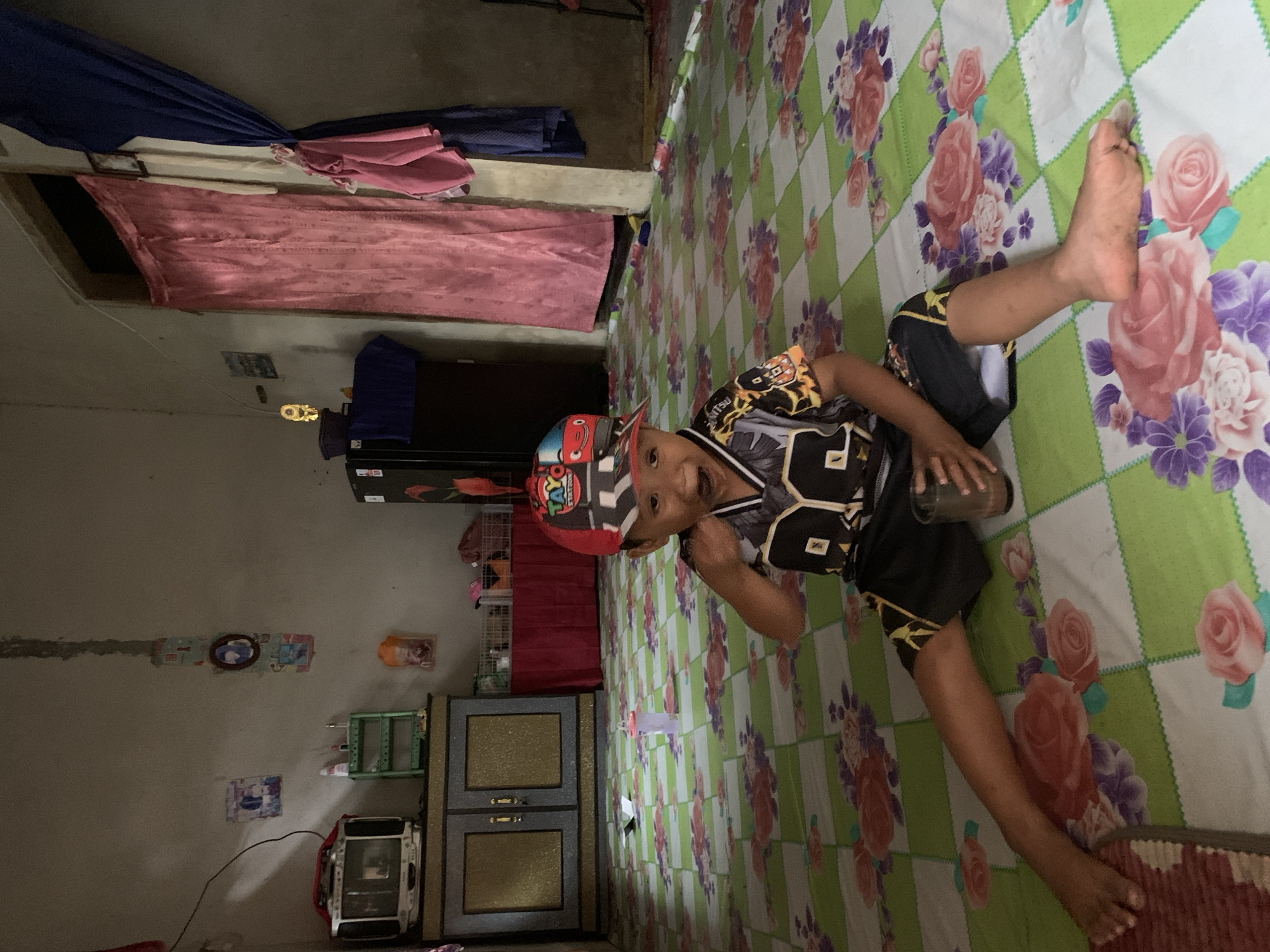 In a dimly lit indoor room, a young child wearing a colorful baseball cap and a black and yellow jersey sits on a floral-patterned mat. The child is holding a glass of brown liquid. To the left, a vintage television set rests on a cabinet with gold accents. Further left, a small green shelf holds various items. In the background, a dark refrigerator is visible, with pink and blue curtains hanging near a doorway. The walls are bare, with a few posters.