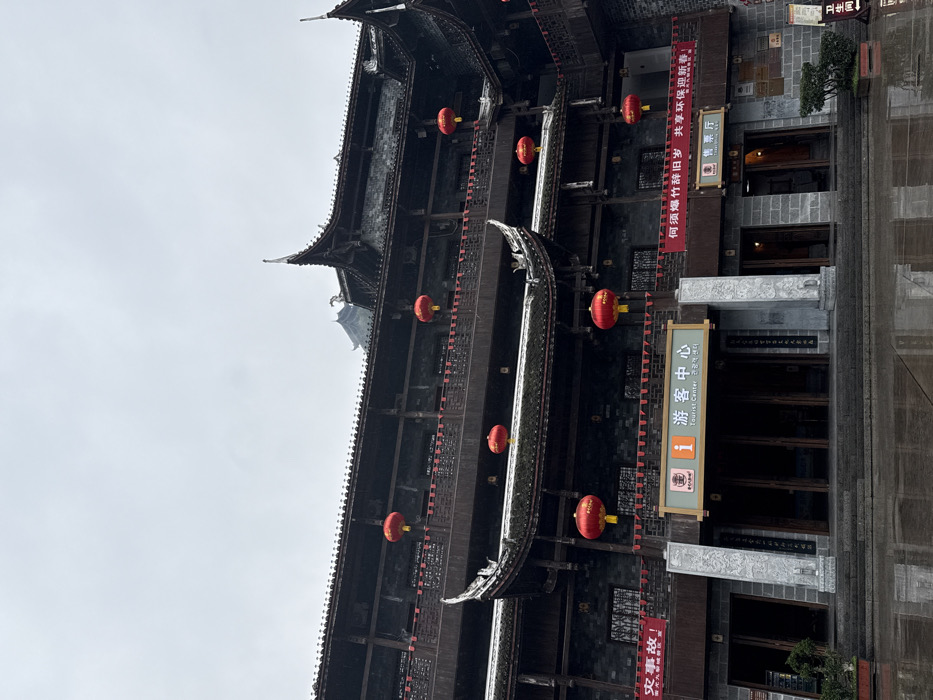 This image displays a multi-story traditional Chinese building, identified as a Tourist Center and Ticket Office, located in Pengshui Miao and Tujia Autonomous County, China. The structure features dark wooden construction with multiple tiered balconies, intricate dark-tiled rooflines with upward-curving eaves, and grey stone elements on the lower facade and pillars flanking the main entrance. Seven spherical red lanterns with yellow tassels are visibly suspended from the balconies and eaves across different levels. Red banners with white Chinese text, including partial messages related to incidents and welcoming the New Year with environmental protection, are strung across the building's facade. The ground in the foreground is dark, wet, and reflective, indicative of recent precipitation. The sky above is uniformly overcast and grey. No individuals are present in the scene. Small potted plants are positioned near the building's entrance.
