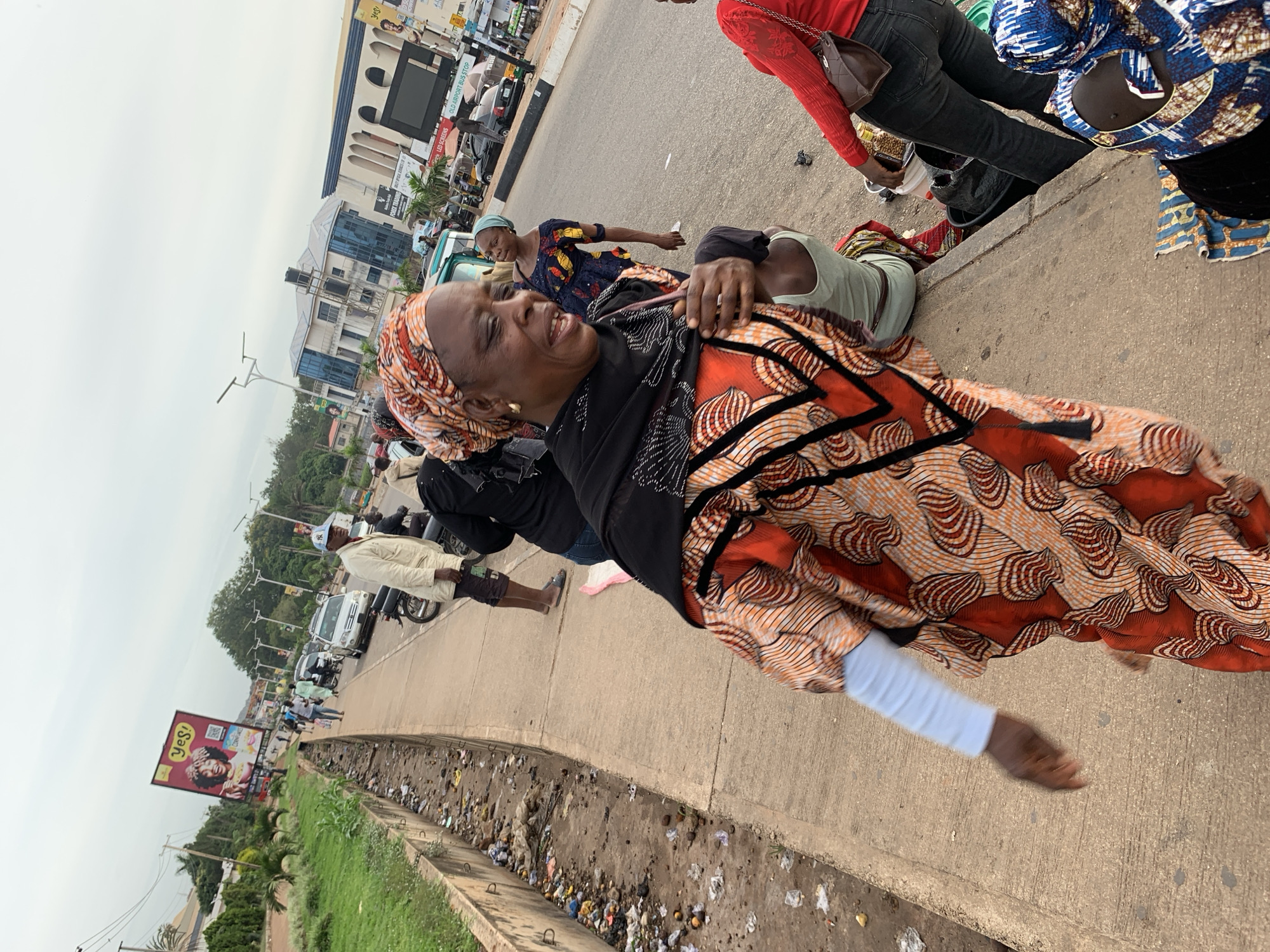 This image depicts a street scene in Barakin Lerawi, Nigeria. In the foreground, a woman wearing a vibrantly patterned orange and white dress and a matching headscarf is the primary subject. She is looking to her right with her mouth slightly open. Behind her, a person in a blue and yellow patterned outfit and headscarf stands. To the right, another woman in a red top and black jeans is visible, holding a brown bag. In the background, various vehicles, including cars and motorcycles, are on the road. A billboard advertising "YES!" with a woman's image is prominent on the left side of the frame. The environment shows a concrete sidewalk alongside a drainage ditch filled with refuse, suggesting an urban setting. The sky is overcast.