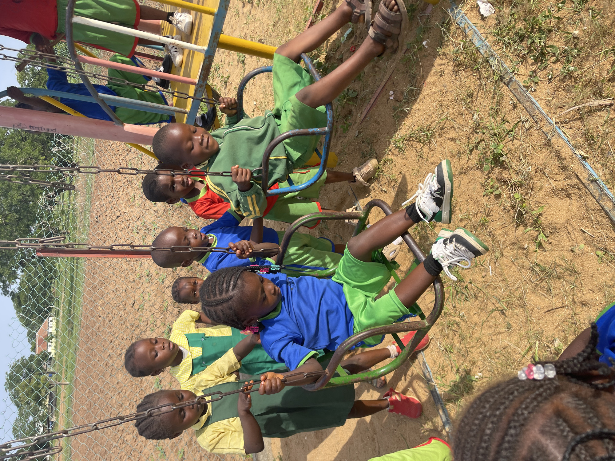 The image shows a group of children on a merry-go-round in what appears to be a schoolyard in Akwanga, Nigeria. The children are of African descent and are wearing school uniforms of various colors: yellow and green, blue and green, and green. They are sitting on the seats of the merry-go-round and holding onto the chains. The merry-go-round is made of metal and has a yellow and blue frame. The ground is dirt and sand with sparse patches of grass. A chain link fence is visible in the background, with a few trees and buildings in the distance. The sky is clear and blue. The image is taken during daylight hours, with the sun casting shadows on the ground.