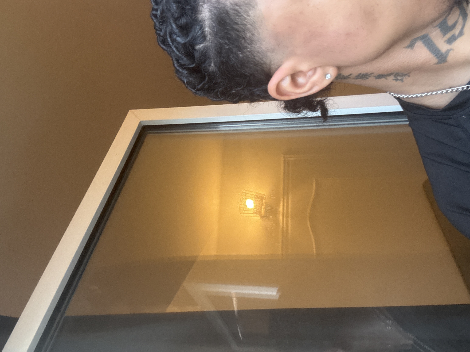 The image shows a close-up view of the right side of a person's head and neck, partially reflected in a glass surface. The person has dark, braided hair and is wearing a silver chain necklace. A tattoo is visible on their neck. The background is an indoor space with a warm, yellowish light, possibly from a lamp or overhead lighting, reflecting on the glass. There is no discernible information in the image to indicate a specific location within the city of Massy, France.