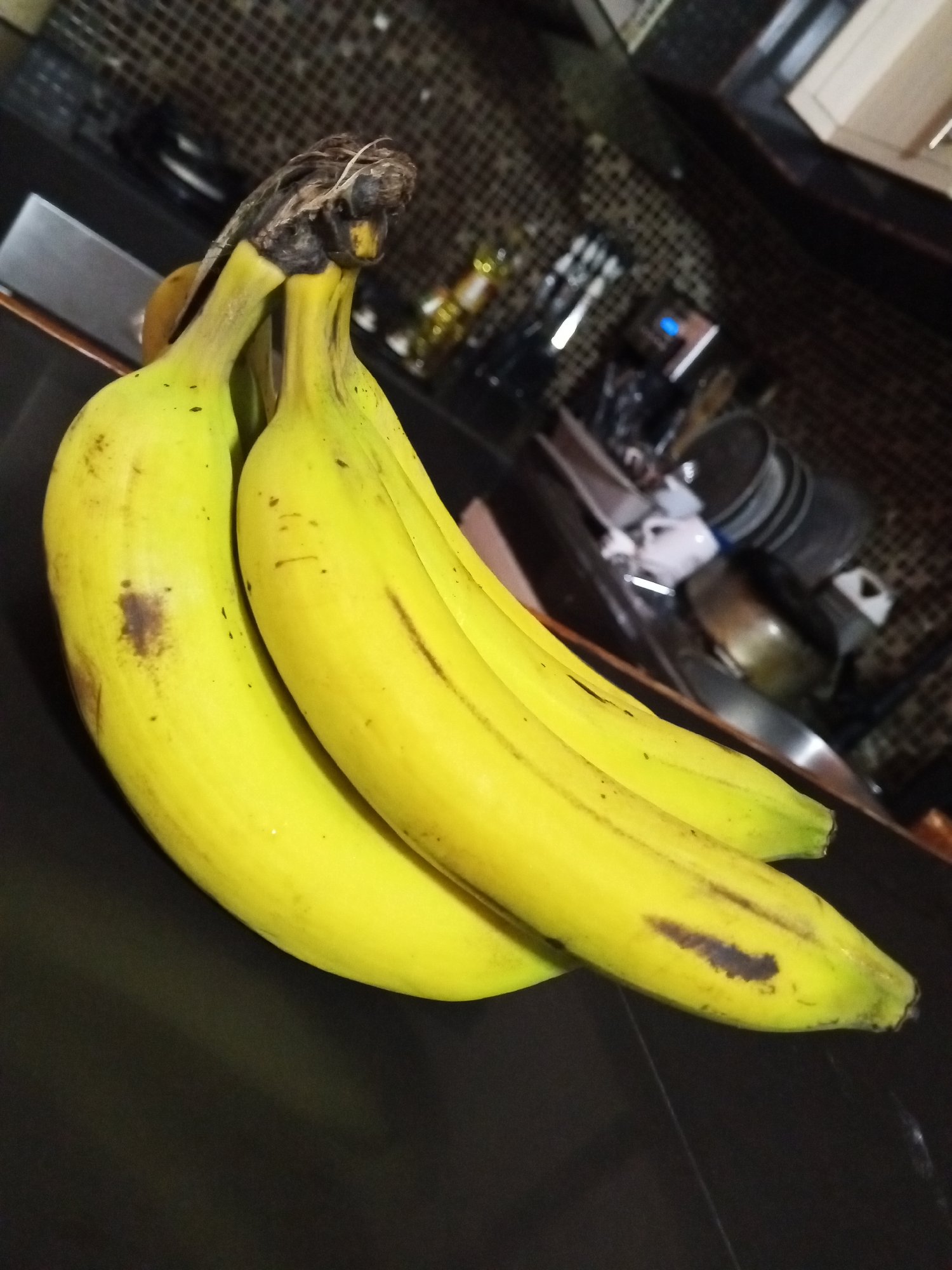 A cluster of three bananas is laying on a dark, possibly black countertop. The bananas are yellow, with some spots of brown discoloration. The stem of the cluster is brown and dry. The background is blurry and features kitchen items such as a stainless steel oven, dishes, and jars. The location is Unknown, Unknown.