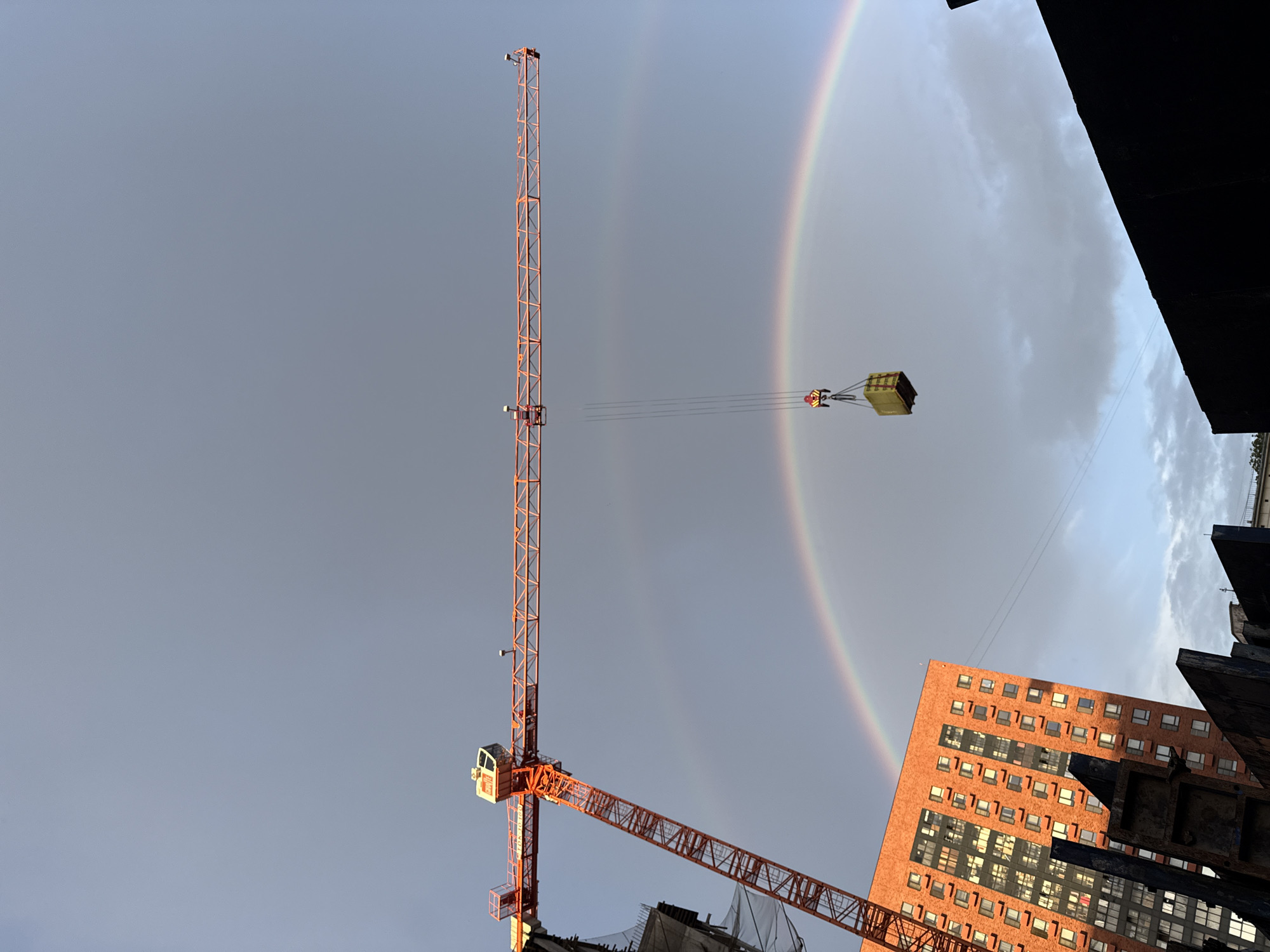 The image depicts a construction site in Moscow, Russia, with a large orange tower crane dominating the frame against a cloudy sky with a double rainbow. A yellow cargo box is suspended from the crane by cables, and a person wearing a safety helmet is visible operating the crane. In the background, a tall, modern brick building with numerous windows is illuminated by sunlight. The foreground shows dark, angular structures, likely part of the construction site.