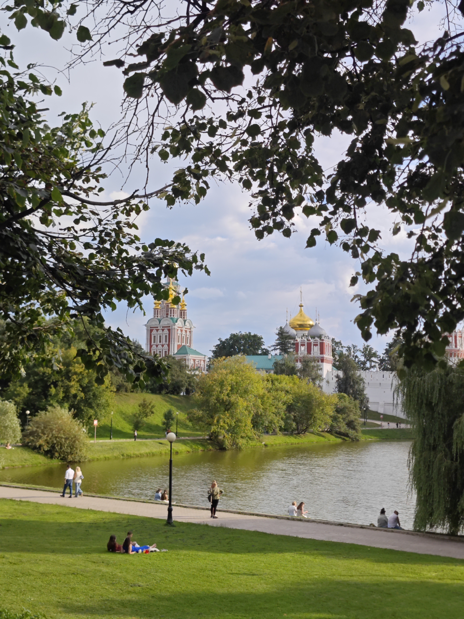 This is an outdoor, eye-level, full shot taken on a sunny day in Moscow, Russia, at Novodevichy Convent. The foreground is dominated by lush green grass with several people scattered across it. In the middle ground, a serene lake reflects the sky and surrounding trees. A paved walkway runs along the edge of the lake, with a few individuals strolling or sitting by the water. In the background, the iconic red and white Novodevichy Convent with its golden domes stands prominently, surrounded by verdant trees. The branches of leafy trees frame the top of the image.