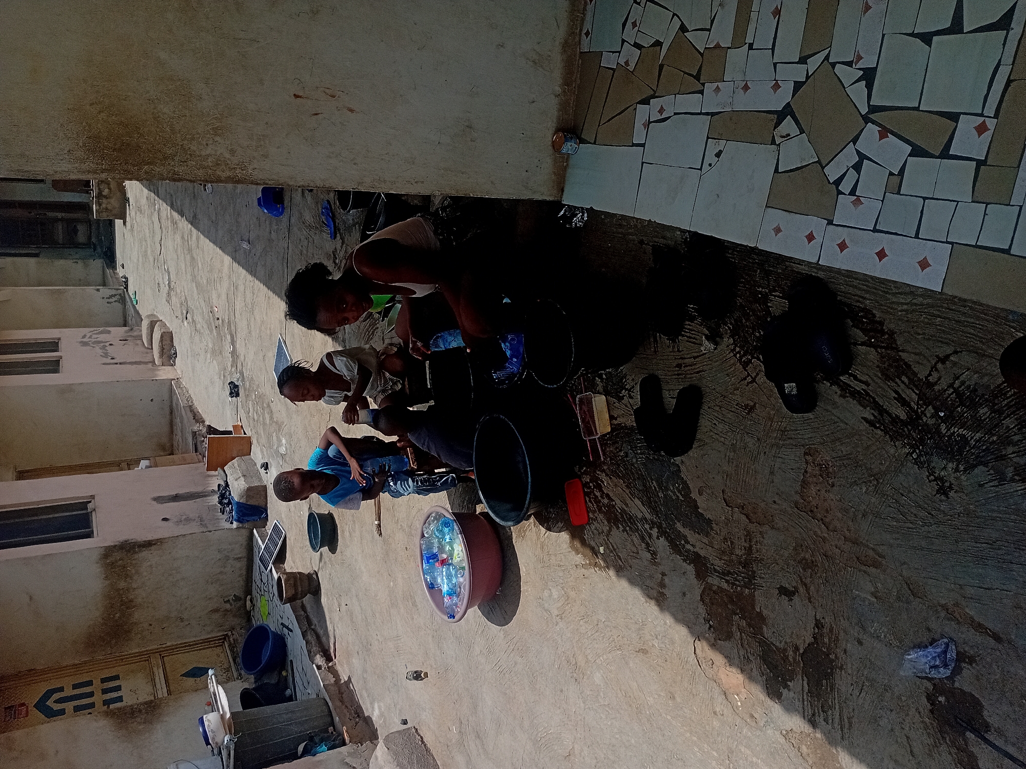 The image captures an outdoor scene, likely a courtyard or common area, with several individuals engaged in activities. In the foreground and mid-ground, three to four people are visible. One person, wearing a white top, is bent over a black bucket, seemingly washing or cleaning. Another individual, dressed in a blue t-shirt and jeans, is seated or squatting nearby, also focused on tasks involving containers. Further back, another person in a green shirt is visible.
The ground is a concrete surface, marked with wet patches and general debris. Scattered across the area are numerous objects, including several plastic buckets (black, blue, and red), a large reddish-brown basin filled with plastic water bottles, and a small solar panel. Other items like sandals/slippers and brooms are also present.
The setting includes buildings in the background, which appear multi-storied with visible windows, suggesting a residential or communal environment. A distinctive feature is a section of the ground or a low wall in the upper right, adorned with a mosaic pattern made from broken ceramic tiles.
The strong, defined shadows cast by the people and objects indicate direct sunlight, placing the scene in the daytime, likely mid-morning or late afternoon. The overall environment, including the architecture and the items present, suggests a warm climate and potentially a developing region. On a wall in the distant background, a blue arrow symbol with some geometric shapes or text below it, possibly "EES," is vaguely discernible. The activity appears to be a communal chore, possibly involving water management or domestic cleaning.