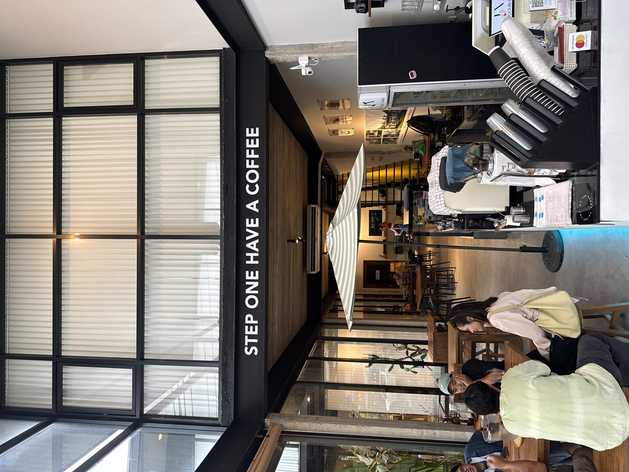 The image depicts the interior of a modern coffee shop, identified as "STEP ONE HAVE A COFFEE" by signage. The establishment is located in the city of Ko Samui District, Thailand. The space features a high ceiling with a black metal frame structure overlaying sheer white blinds on the upper windows, and large glass windows and doors allowing natural light to enter.
Inside, several patrons are seated at wooden tables, some engaged in conversation. In the foreground, a group of three individuals is seen at a table. One person, wearing a light green tie-dye t-shirt, is facing away from the camera, while another, in a pink long-sleeved shirt, is facing them. A third person, wearing a light blue cap, is also at the table, looking towards the individual in the pink shirt.
The coffee shop appears to be well-equipped, with a counter area visible in the mid-ground. This counter includes a coffee machine, a display of cups, and a POS system. A refrigerator unit is also present. An outdoor umbrella with a striped pattern is positioned outside, providing shade. Artwork or decorative items are displayed on the walls, and plants are visible, adding to the ambiance. A security camera is mounted on the wall near the ceiling. The overall atmosphere suggests a casual and contemporary setting for patrons to enjoy coffee.