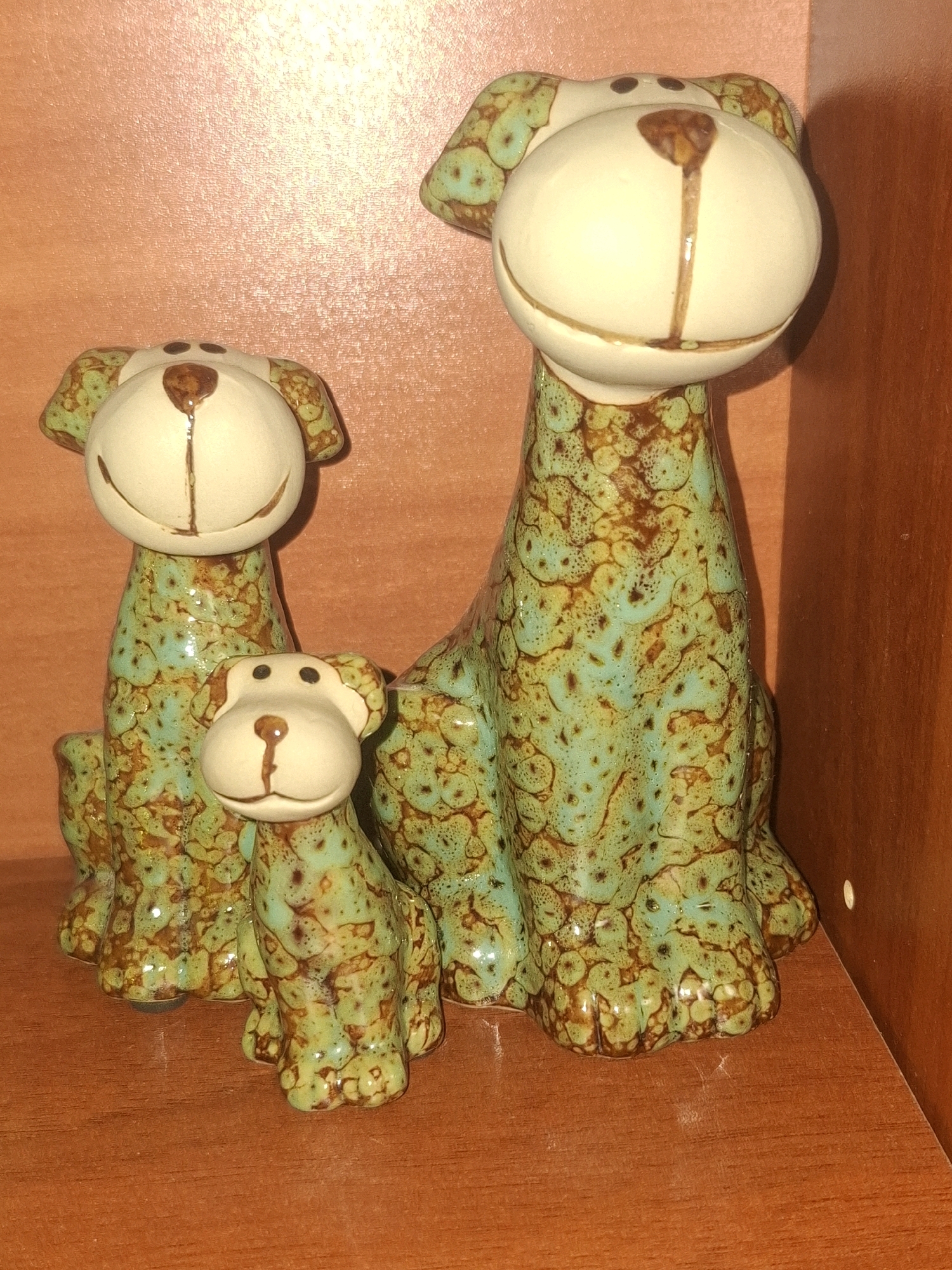 Three ceramic dog figurines are displayed on a wooden shelf. The figurines are a pale cream color with a speckled green and brown glaze. The largest figurine is in the back right, with a medium-sized figurine behind it to the left. The smallest figurine is in the front, centered between the other two. The background is a textured, peachy-pink wall, and to the right is a vertical wooden surface, possibly part of a cabinet or shelf unit. The lighting is warm and appears to be coming from the front and slightly to the left, casting subtle shadows. This scene is not specific to Almaty, Kazakhstan, and could be found in any domestic setting.
