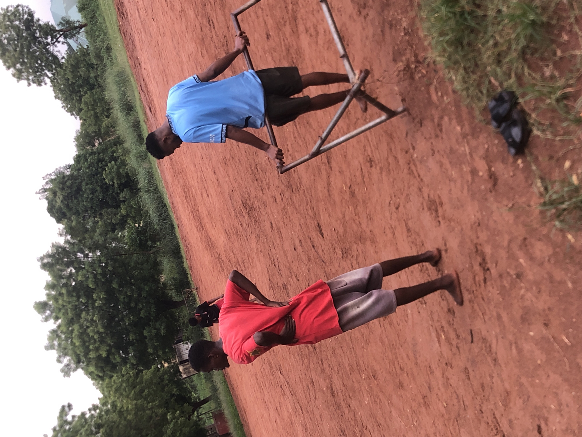 This outdoor image, rotated 90 degrees clockwise, captures two young males on a reddish-brown dirt field. The field, likely a playground or open area, is characterized by its earthy tone and some scattered patches of green grass along the edges. Lush green trees and dense foliage form the background, suggesting a natural or semi-rural setting under an overcast or hazy sky.
In the foreground, a young male wearing a bright red t-shirt and light grey shorts stands facing away from the camera, looking towards the right side of the frame. His hands are placed on his hips or lower back, and he appears to be observing or taking a break. Further back and to the right, another young male, dressed in a light blue t-shirt with patterned sleeves and dark shorts, is engaged with a metal frame structure. He is holding onto the upper bars with both hands and has his feet placed on a lower crossbar, suggesting he is using it for exercise, like dips, or simply balancing on it. The metal frame appears to be a makeshift piece of equipment or a goalpost.
To the right of the person in blue, a dark, indistinct object, possibly a bag or folded clothing, rests on the ground. The overall lighting suggests it might be late afternoon or early morning, with soft, diffused light and no harsh shadows. The reddish soil and tropical vegetation are consistent with the provided location context of Ukwu-Abwa, Nigeria. There is no visible text in the image.