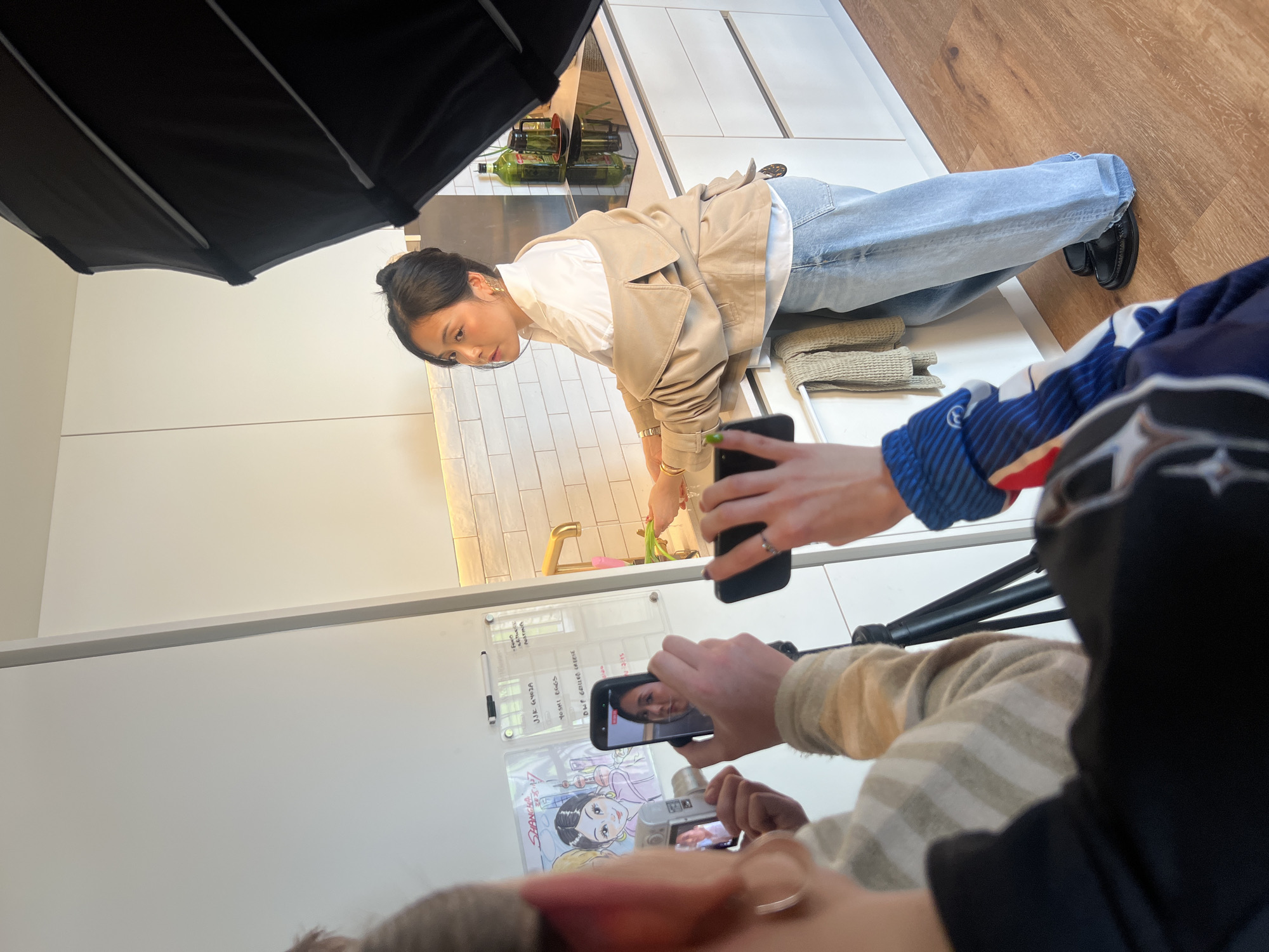 A woman is bending over a kitchen sink, possibly washing dishes, as two people photograph her with smartphones. She is wearing a white shirt, a light tan jacket, and light blue jeans, and black loafers. The kitchen has white cabinets and white tiled backsplash. One photographer is wearing a striped top, and the other is wearing a blue and white sports-themed top. Behind the woman are shelves with cooking oils. A lighting umbrella is visible above her, and a tripod is visible between the photographers. A whiteboard with writing and a drawing are visible on the left. The location is likely a studio set rather than a real kitchen in London.