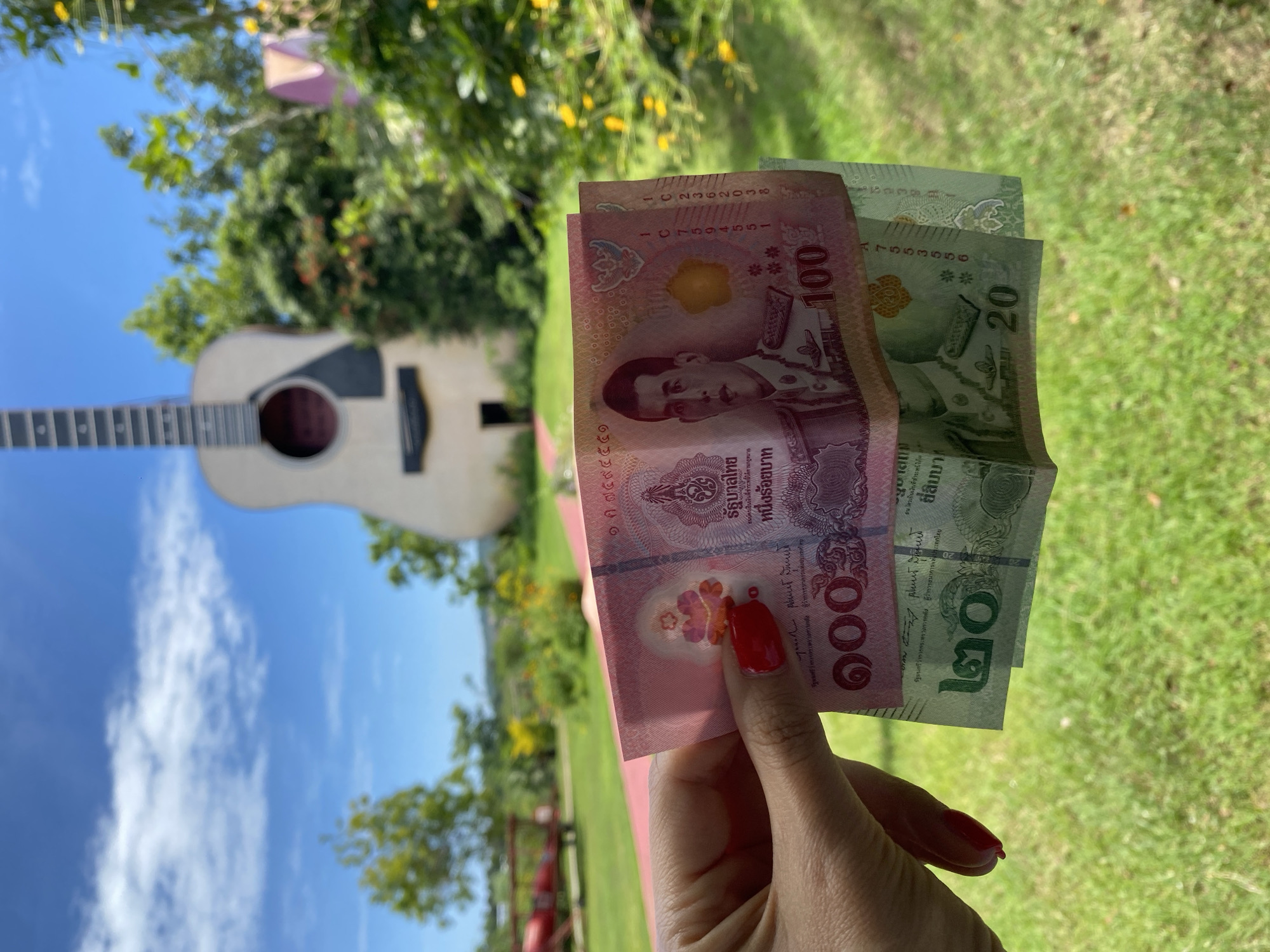A person's hand with red nail polish holds two Thai baht banknotes. The top banknote is pink and features a portrait of a king, along with the number 100. The bottom banknote is green and shows the number 20. The banknotes are held in front of a blurry outdoor background. In the background, a very large, light-colored acoustic guitar structure stands prominently. Beyond the guitar, there are green trees, yellow flowers, and a blue sky with scattered clouds. To the left, a red structure resembling a small plane is visible. The scene is set in Bang Sare, Thailand.