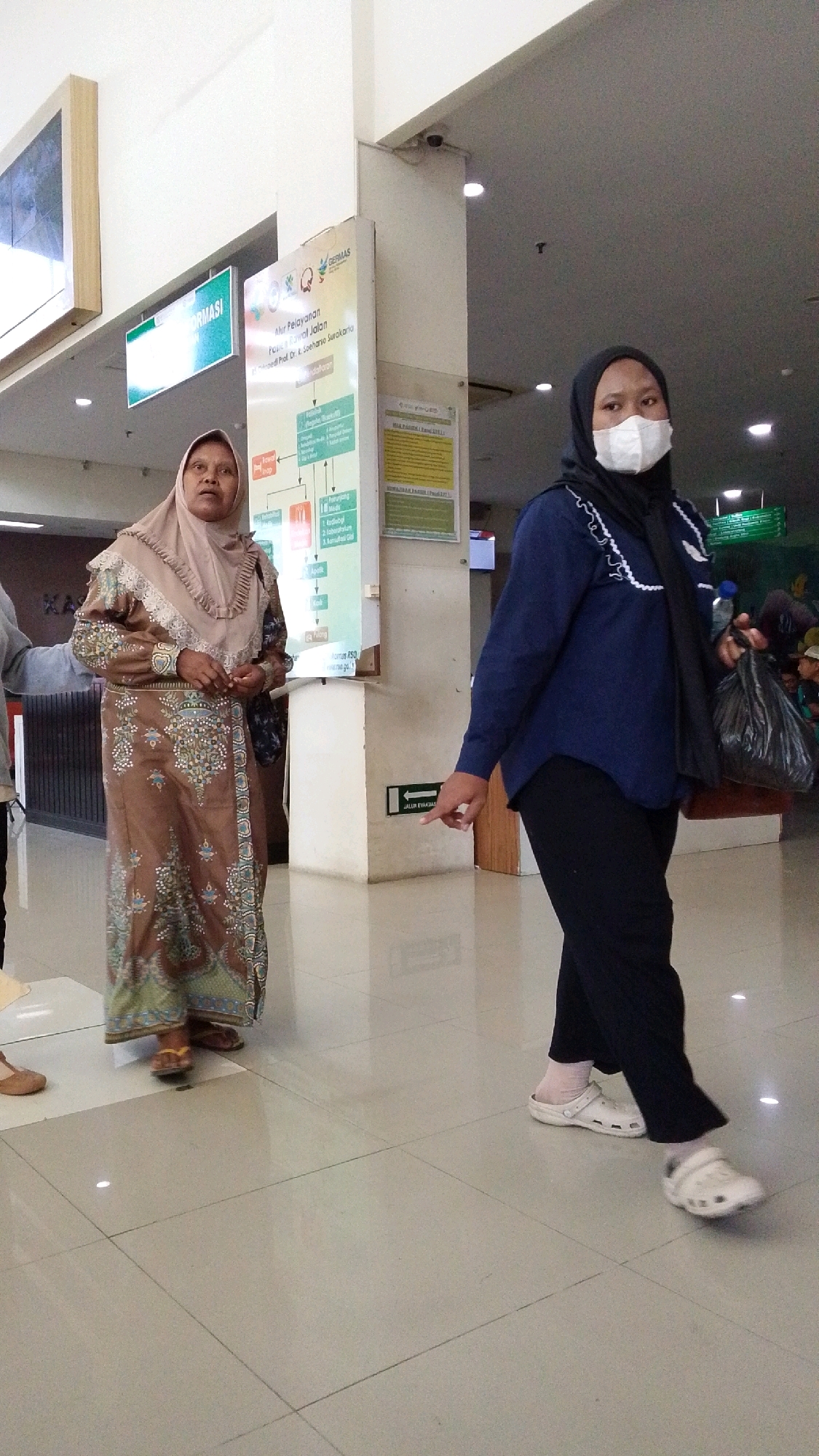 Two women are walking in a well-lit indoor space, possibly a hospital or clinic lobby, in Pabelan, Indonesia. The woman on the left is wearing a beige hijab and a long, brown, patterned dress with a ruffled collar and sleeves. She is looking to her left. The woman on the right is wearing a black hijab and a white face mask. She has on a dark blue long-sleeved shirt, black pants, white socks, and white clogs. She is carrying a black plastic bag in her right hand and looking forward. In the background, there are information boards, signage, and what appears to be a security camera on the ceiling. The floor is tiled in a light color with visible grout lines, and there are reflections from the overhead lighting.