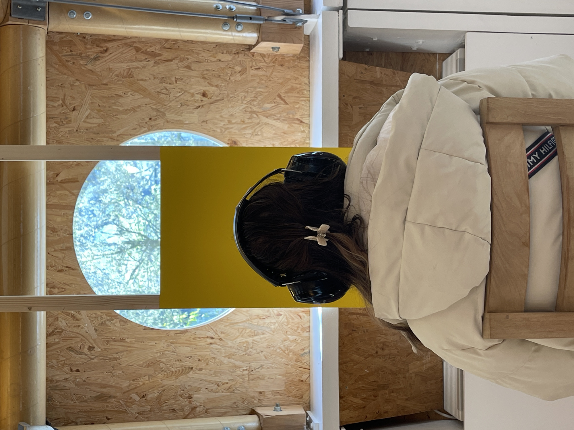 A person wearing headphones stands in front of a structure made of wood and metal, possibly inside an architectural installation in Metz, France. The structure features a round window providing a view of trees and sky, and is partially painted yellow. The person is wearing a light-colored jacket and a white sweater with a visible "Tommy Hilfiger" logo on the collar.