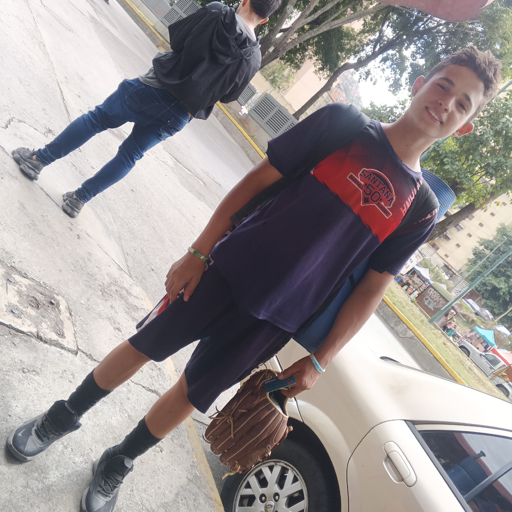 The image captures two young males in an outdoor urban setting in Caracas, Venezuela, during the daytime. In the foreground, a smiling young male, likely a teenager, looks directly at the camera. He has short, dark, curly hair and is dressed in a dark blue t-shirt featuring a red and black design on the chest that reads "SANTANA BASEBALL 50 ACADEMY." He is also wearing dark shorts, black socks, and dark grey sneakers. In his left hand, he holds a brown "Rawlings" baseball glove along with a small blue card. A blue backpack strap is visible over his right shoulder, and he wears green and white, and light blue wristbands.
Behind him, a second young male is visible from the back, walking away. He is wearing a black hooded jacket, blue jeans, and dark grey sneakers.
The setting is a concrete sidewalk next to a street, demarcated by a yellow curb and a grassy strip with trees. In the background, multi-story buildings are visible, along with what appear to be street vendor stalls featuring colorful canopies (blue and red), suggesting a bustling street scene. A light-colored car is partially parked on the right side of the frame. The overall impression is a casual, sunny day, with the main subject seemingly involved in a baseball-related activity.