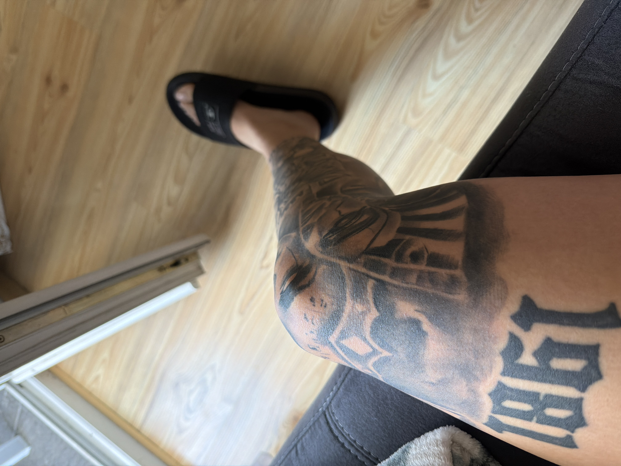 The image shows a close-up view of a person's tattooed leg, resting on a dark fabric surface, likely a sofa. The leg displays extensive black ink tattoos, including the numbers "1861" prominently on the lower part of the thigh. Other tattoo elements, appearing to be artistic designs, are visible on the knee and upper thigh. The individual is wearing a black slide sandal on their visible foot. The background consists of a light-colored wooden floor and a section of a white door frame. There are no discernable elements in the image that would allow for the determination of its location within the city of Plovdiv, Bulgaria.