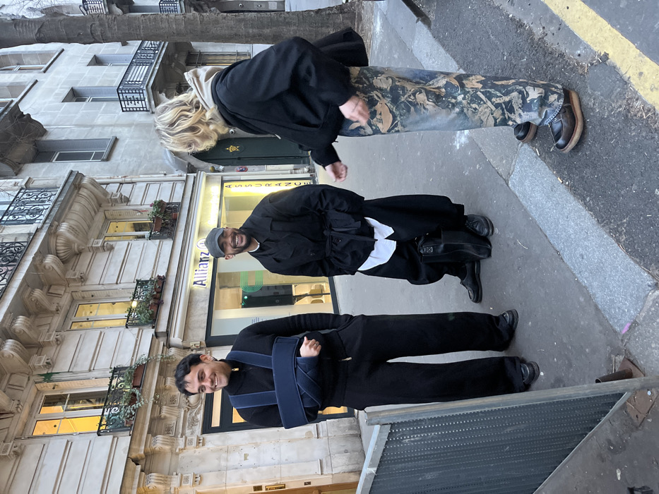 Three people stand on a city sidewalk. One person with blonde hair wears a black jacket and patterned pants. Another person in the middle wears a black coat and a gray hat. The third person at the bottom wears a black top and black pants with a blue arm sling. Buildings with ornate balconies line the background. The sidewalk is adjacent to a street with a yellow line. An "Allianz" sign is visible on a building.
