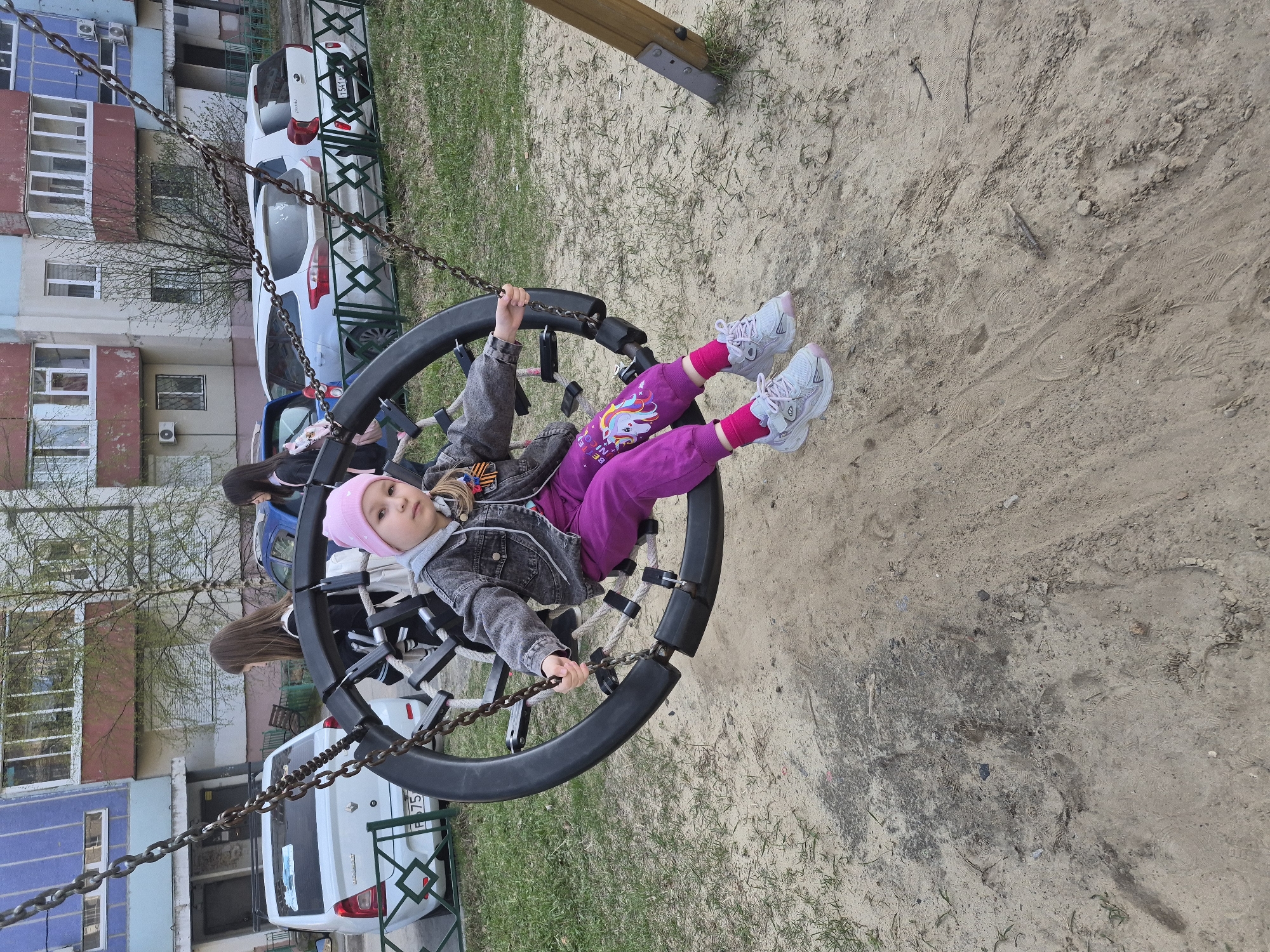 A young girl, likely between 6 and 8 years old, is the central subject of the image. She is wearing a pink beanie, a gray denim jacket over a hooded sweatshirt, pink pants with a unicorn graphic, and white sneakers. The girl is seated inside a circular swing, suspended by chains from a wooden structure. She is holding the swing's chains with both hands.
The location appears to be a playground in an urban setting. In the background are multi-story apartment buildings with varied architectural details, indicating a residential area. Several parked cars are visible near the buildings, and some green vegetation suggests the presence of trees or landscaping. The ground surface beneath and around the swing is primarily sand.
While the photograph was taken in Russia, the image does not contain information that would confirm its location in Tobolsk.