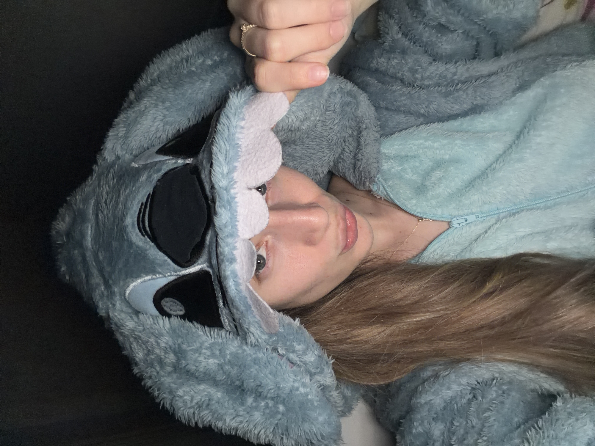 A close-up, angled shot features a person, likely female, lying down indoors, wearing a cozy, light blue and grey fuzzy onesie that resembles the character Stitch from Lilo & Stitch. The person has fair skin, light-colored eyes, and long, wavy brown hair that cascades beside their head. Their lips appear glossy, and a delicate gold-colored ring is visible on their index finger, which is partially obscuring their face. A thin necklace chain is also visible near the zipper of the onesie.
The onesie features distinct embroidered elements of Stitch's face on the hood, including large black and white eyes and the inner ear details in light pinkish-purple and lighter blue. The fabric of the onesie is plush and soft, suggesting comfort. The background is mostly dark and out of focus, indicating an indoor setting, possibly a bed or couch, creating a snug atmosphere. The overall impression is one of relaxation and comfort. No text is visible in the image. The location context provided is Plovdiv, Bulgaria.