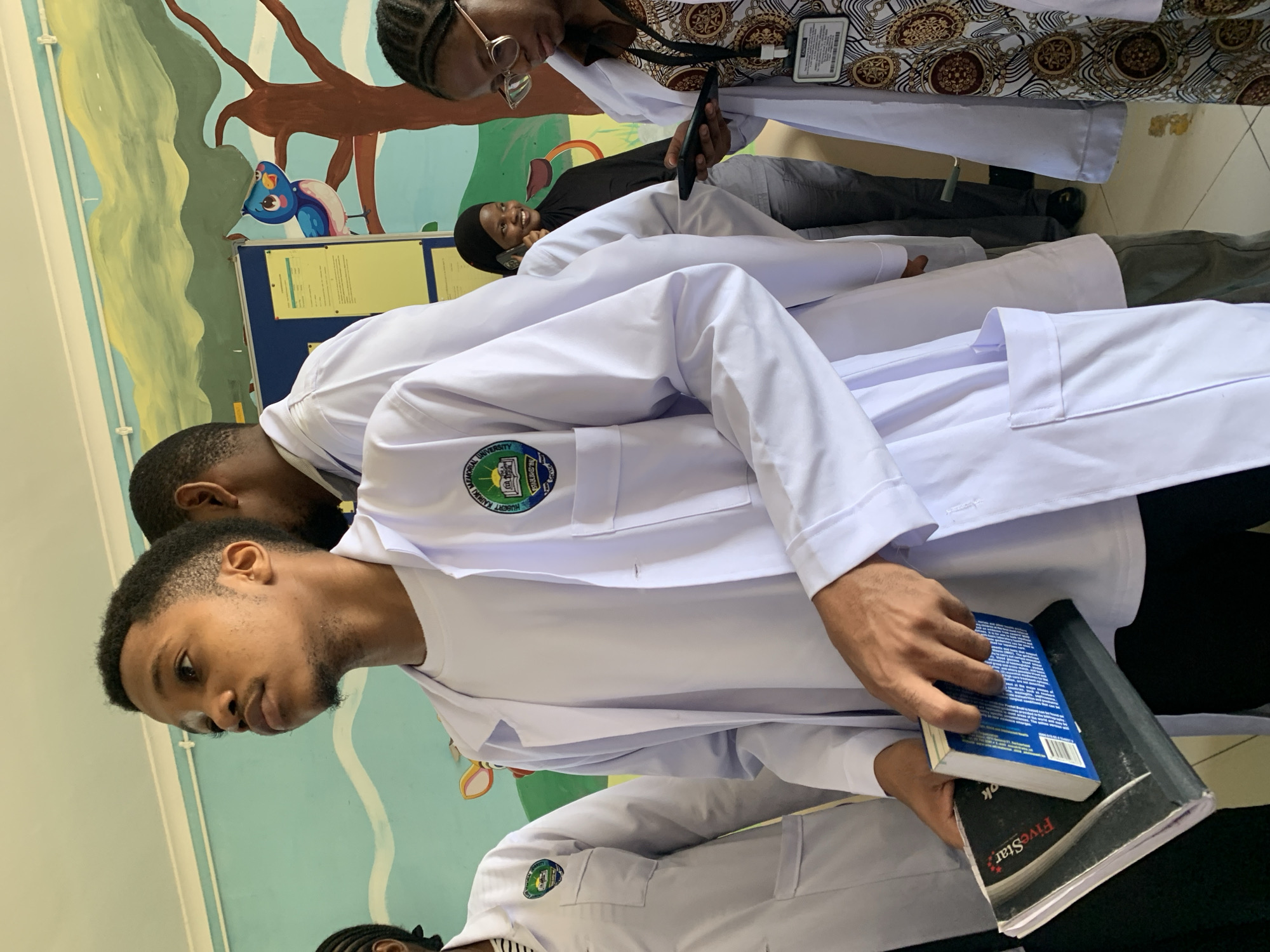 An indoor photograph captures a group of young adults, likely students at Hubert Kairuki Memorial University in Dar es-Salaam, Tanzania, dressed in white lab coats. The setting appears to be a brightly lit hallway or common area within the university.
In the foreground, a young man with a beard is holding a blue book and a black notebook labeled "FiveStar," looking thoughtfully downwards. Behind him and to his right, another student, wearing glasses and a patterned top over a lab coat, holds a mobile phone. Further back, an individual in a black hijab and gray trousers is visible, smiling faintly while talking on a phone. Several other individuals in white lab coats are partially visible throughout the scene.
The background features a bulletin board with various documents and a colorful wall mural depicting trees and a whimsical blue bird, suggesting a vibrant educational environment. The general mood is calm, indicating a casual moment between classes or activities during the daytime. The visible logo on the lab coats clearly displays "HUBERT KAIRUKI MEMORIAL UNIVERSITY," confirming the institutional context.