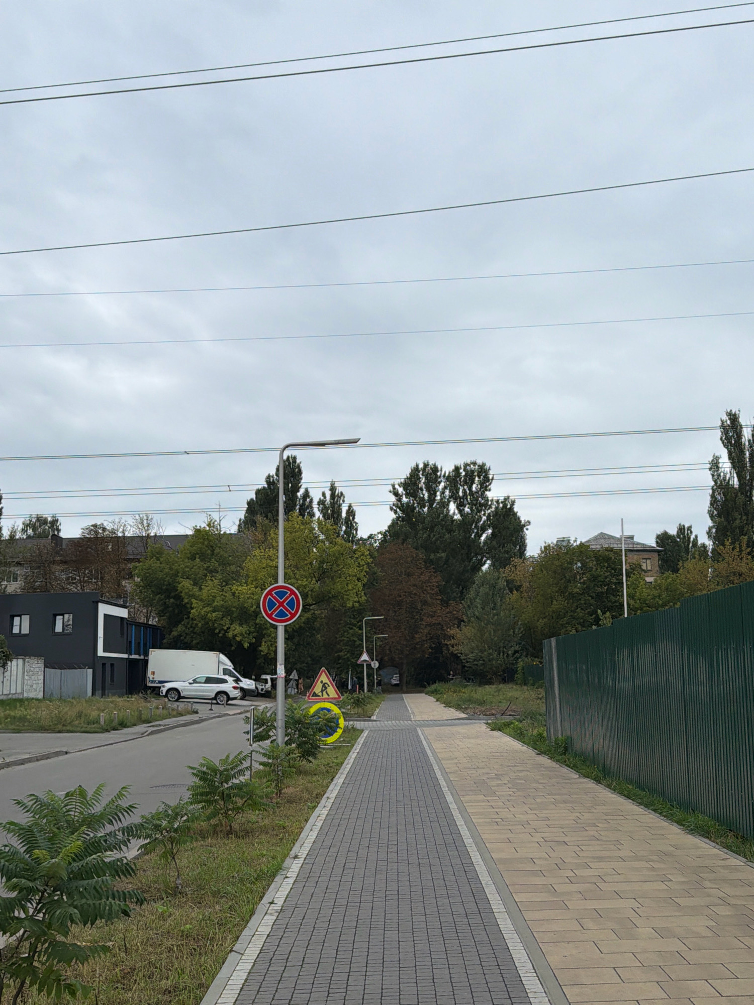 The image shows a street scene in Kyiv, Ukraine. On the left side of the street, there is a modern building with dark gray and white facades. A white van and a silver car are parked on the side of the road. A traffic sign indicating no parking is visible on a pole. Further down the street, there are trees and more buildings in the background.
The right side of the image features a pedestrian walkway paved with gray and beige tiles. A tall green metal fence runs along the edge of the walkway. In the middle ground, there is a yellow triangular traffic sign depicting a person walking, indicating a pedestrian crossing. A circular yellow sign with a blue symbol is placed near the pedestrian crossing sign. The sky is overcast with gray clouds. Overhead, multiple power lines are stretched across the frame.