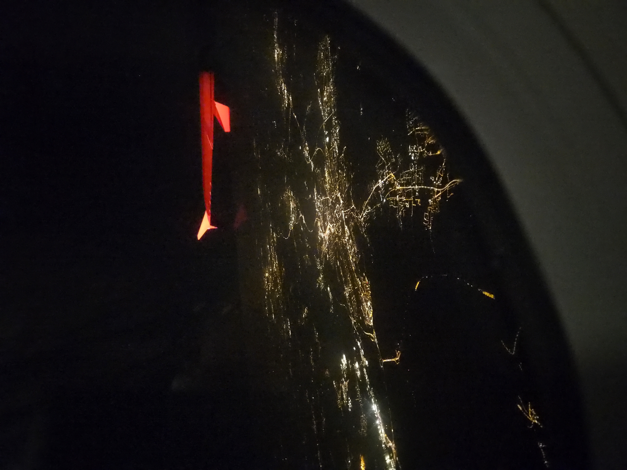 An aerial nighttime view from an airplane window shows a brightly lit city spread out below, with the wing of the aircraft illuminated in red extending into the frame from the top right. The city lights form a dense network of illuminated streets and buildings, suggesting a populated urban area. The lights are predominantly yellow and white, with some appearing as continuous lines representing major roads or highways. The overall impression is of a sprawling metropolitan landscape viewed from a high altitude. The specific location of Rollingen, Luxembourg, cannot be definitively identified from the visual information alone, as the image lacks any discernible landmarks or identifying features.