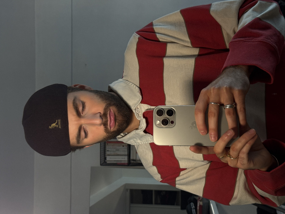 A male subject is depicted in a mirror selfie, visible from the chest up. The individual, estimated to be in his late 20s to early 30s, possesses dark, trimmed facial hair, including a beard and mustache. He is wearing a black Kangol beret with a gold kangaroo logo and a long-sleeved polo shirt featuring wide horizontal stripes in red and off-white, with an off-white collar. His eyes are directed downwards, and his lips are pressed into a pursed expression. Both hands hold a silver-colored smartphone, identified as an iPhone by its triple-lens rear camera module and partial Apple logo reflection, positioned to capture the reflection. On his left hand, a thin gold-colored ring is on the pinky finger and another on the ring finger. On his right hand, a thin silver-colored band is on the pinky finger, and two stacked silver-colored bands are on the ring finger. The background reveals an indoor setting with a light-colored wall. A dark, open shelving unit, containing multiple items consistent with books, is partially visible behind the subject's left shoulder. The image provides no visual cues to ascertain a specific location within Paris, France.