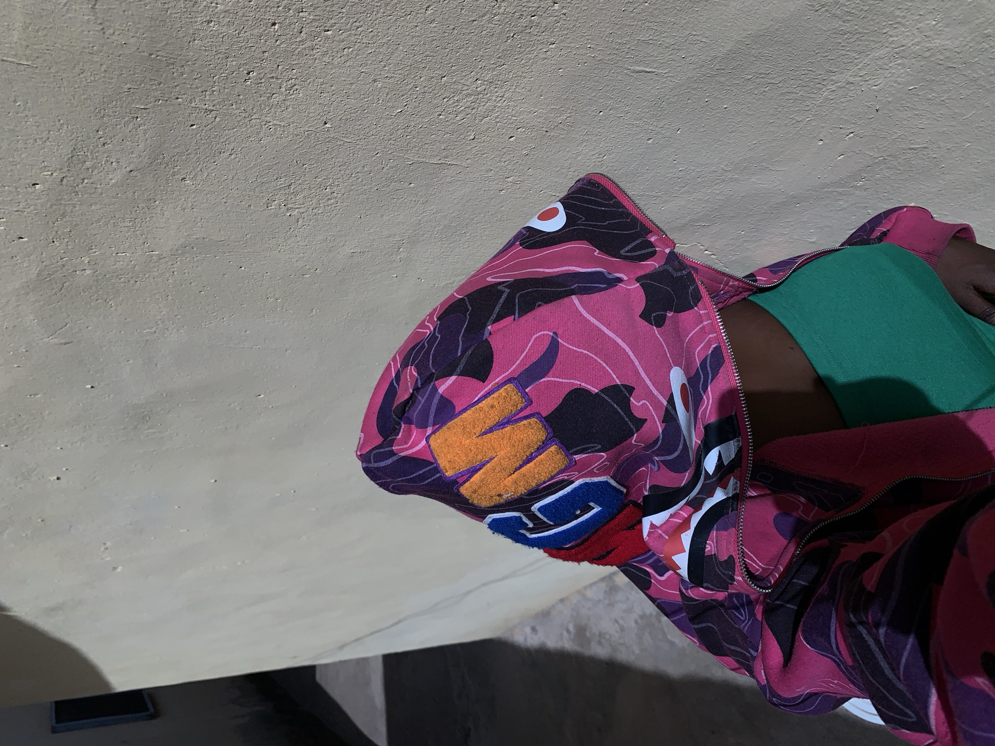A person wearing a pink and black camo-patterned hoodie with the letters "BAPE" on the hood and a green shirt is lying on a surface. The surface appears to be a beige concrete wall. The location is Rafin Par, Nigeria. The image is rotated counter-clockwise.