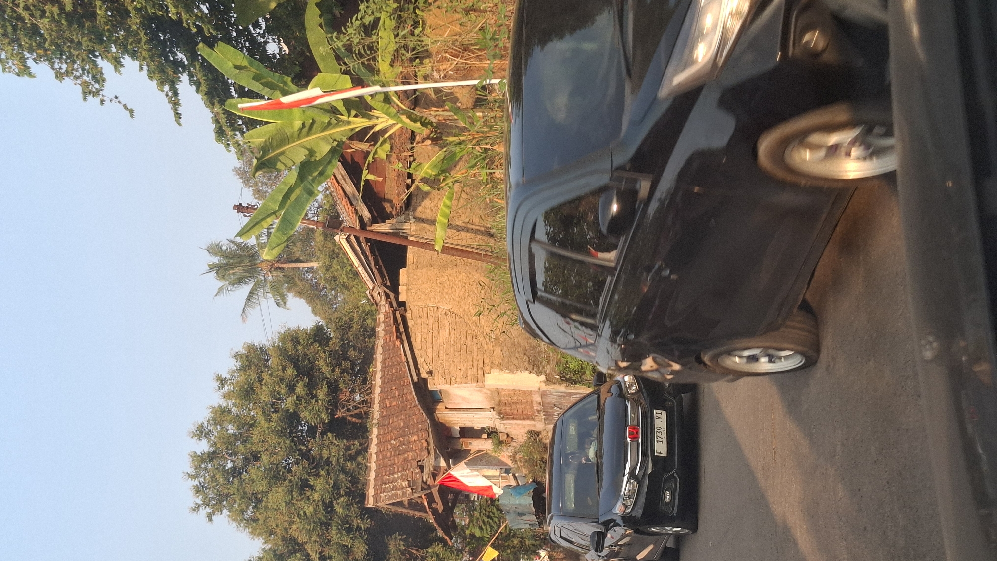 In the city of Sirnagalih, Indonesia, a road scene unfolds under a clear sky with dappled sunlight. Two black cars are the primary subjects. The car in the foreground, partially obscuring the view, is moving. The second car, a black hatchback, is stationary on the left side of the road, displaying a license plate that reads "F 1739 YI". Behind the cars, an aged, rustic house with a tiled roof is visible, with a red and white Indonesian flag displayed prominently on its facade. Lush green trees and banana plants surround the structure, suggesting a rural or semi-rural setting. The overall impression is one of a quiet street with everyday life unfolding amidst the natural beauty of the Indonesian landscape.