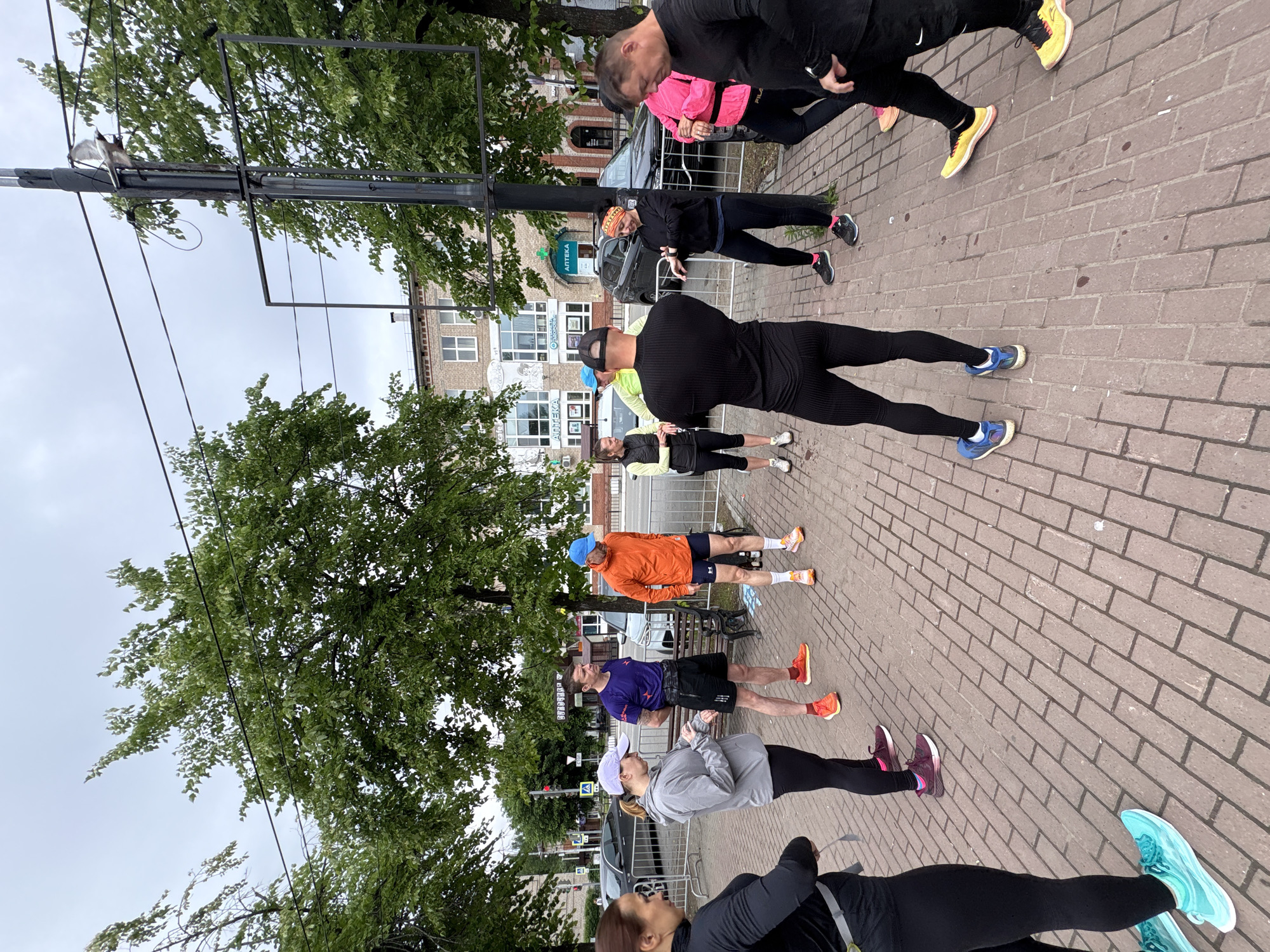 A group of people is stretching on a brick sidewalk in what appears to be the city of Chaikovsky, Russia. The group consists of nine individuals, all dressed in athletic attire. They are lined up in a row, performing various stretching exercises. The street setting is marked by trees, light poles, and buildings. The Russian word "Аптека" (Pharmacy) on one of the buildings provides further support that the scene is indeed in Russia.