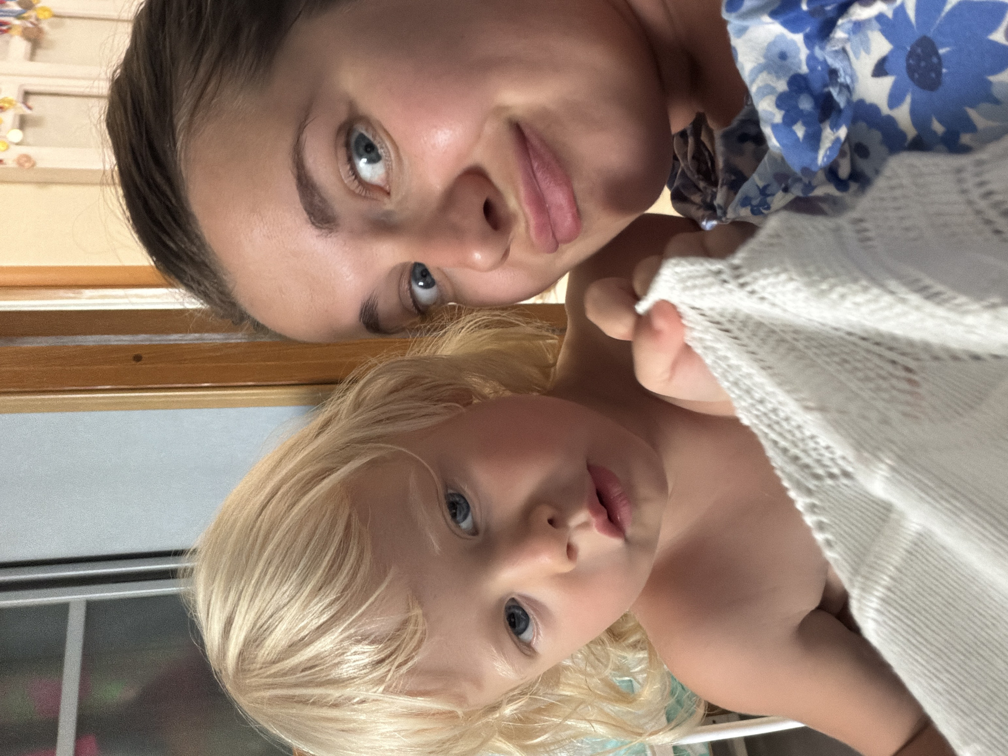 The image shows a close-up, selfie-style shot of two people, an adult and a child, with their faces positioned upside down relative to the viewer. The adult, with dark hair and blue eyes, is looking directly at the camera. They are wearing a blue and white floral top. To their right, a child with blonde hair and blue eyes is also looking towards the camera. The child appears to be peeking out from under a white textured fabric. The background suggests an indoor setting, possibly a home, with wooden elements and what looks like a window with blinds. The lighting suggests it could be daytime. The overall mood appears to be intimate and casual, perhaps a moment captured between a parent and child. There is no visible text in the image. The context of Sochi, Russia is provided, but there are no specific visual cues within the image to confirm this location.