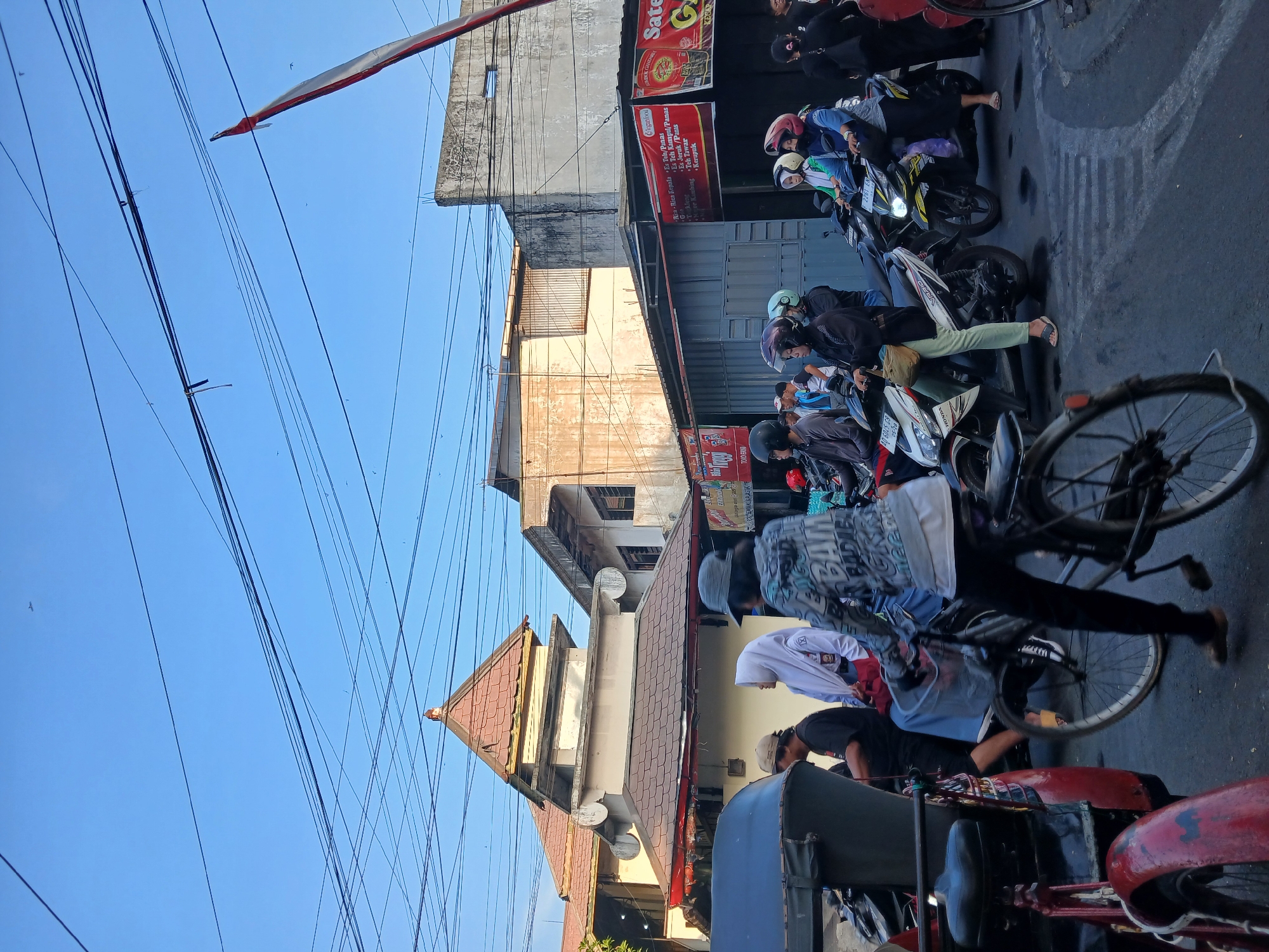 The image captures a street scene in Surakarta, Indonesia, during daylight hours. A traffic jam is evident, with numerous motorcycles and a trishaw forming the primary modes of transportation. People are dressed in casual clothing, with some wearing helmets, indicating road safety awareness. The architecture of the buildings suggests a mix of traditional and modern styles, with a prominent mosque-like structure featuring a tiered roof. Power lines crisscross the sky, a common sight in urban Indonesian landscapes. Signs in Indonesian language are visible on some buildings, likely indicating shops or businesses. The overall atmosphere appears to be that of a bustling urban environment, with everyday life unfolding on the street.