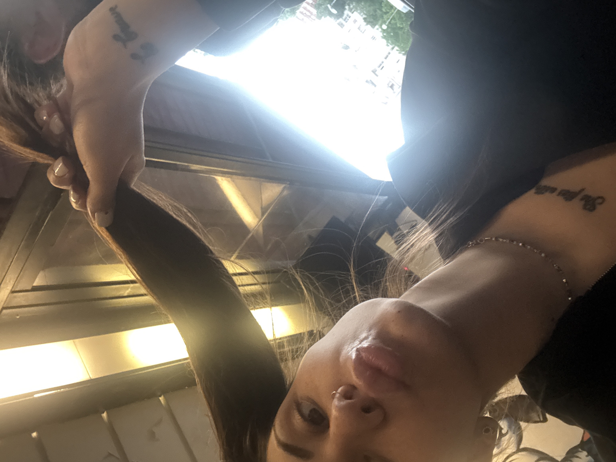 A selfie of a woman inside a vehicle (possibly a bus or train), possibly in Sofia, Bulgaria. The woman's face is prominent, showing details like her nose ring and lips. She is holding a strand of her dark hair. A tattoo is visible on each of her forearms. The background is blurred with bright lights suggesting an interior space with windows. Exterior buildings are partially visible through a window, suggesting an urban environment. The presence of public transportation infrastructure suggests that the location is Sofia, Bulgaria.