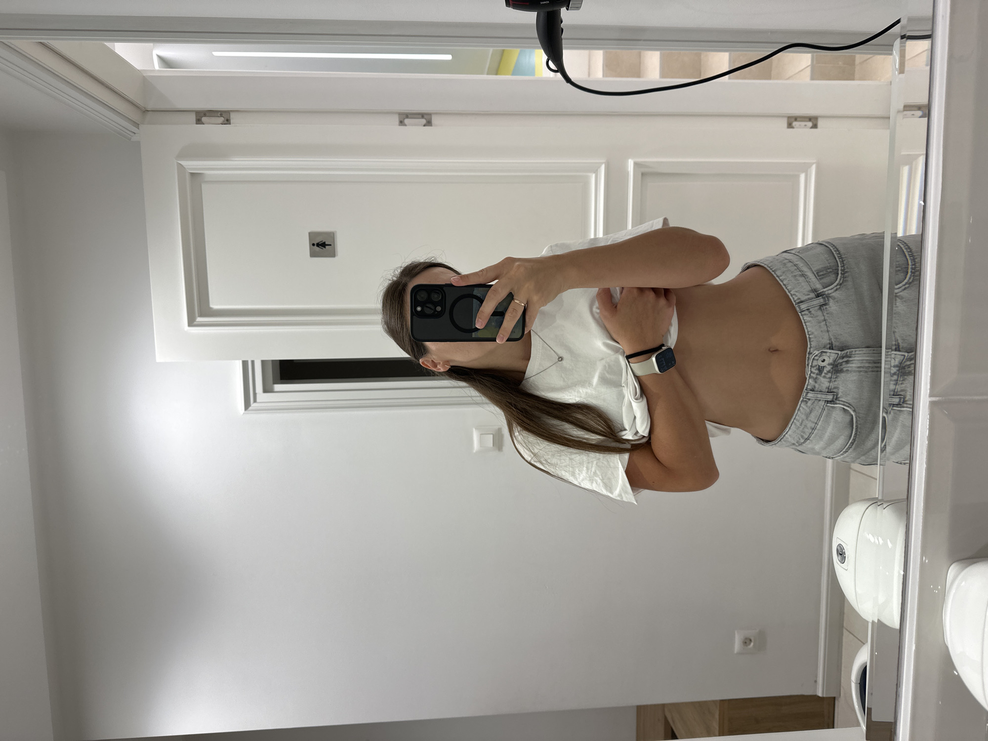 A young woman with long, brown hair is taking a selfie in a mirror. She is wearing a white t-shirt and light-wash denim shorts. Her t-shirt is slightly pulled up, revealing her midriff. She is also wearing a necklace and a smartwatch. The background appears to be a restroom, with a white door and a sign with a female silhouette visible. There is also a hand dryer mounted on the wall. The mirror reflects a sink and a toilet in the foreground. The overall environment is clean and modern. The location is identified as Bečići, Montenegro.