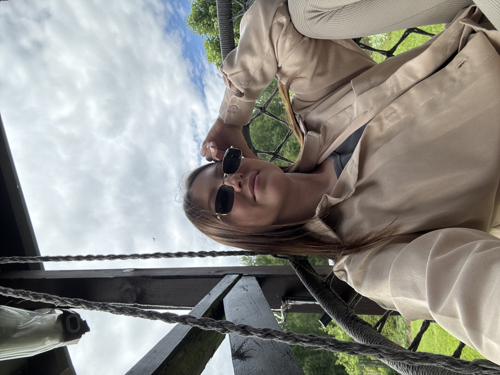 A young woman with long brown hair and sunglasses is sitting in a hanging chair outdoors. She is wearing a tan button-down shirt and light-colored pants. Her left arm is resting on her forehead, and she is looking at the camera. The background shows trees and a cloudy sky. The hanging chair is suspended from a wooden structure. This scene is likely a residential area in the town of Ponaryevo, Russia.