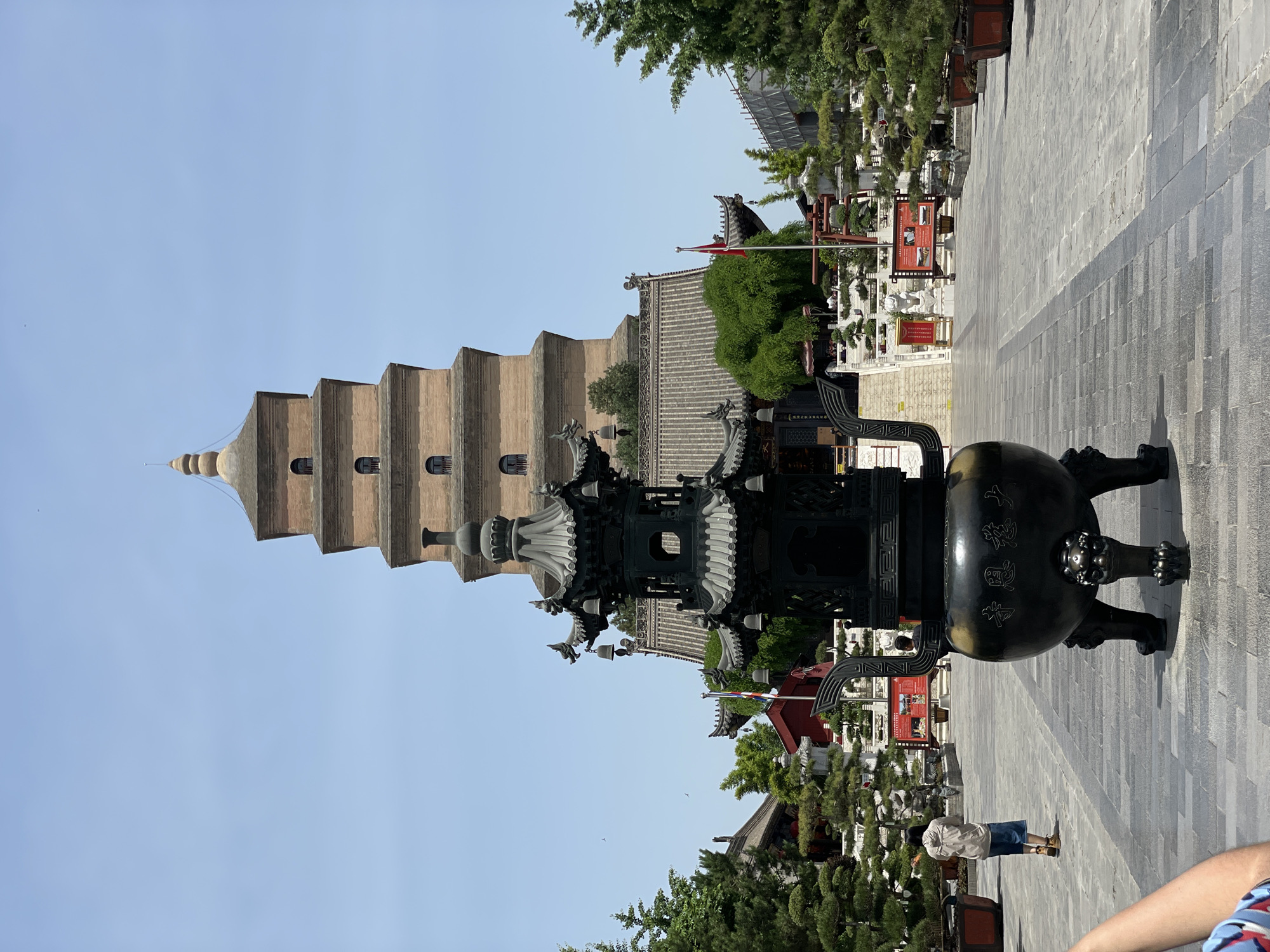 The image shows the Great Wild Goose Pagoda in Yanta District, Xi'an, China. A large bronze incense burner stands in the foreground, with the pagoda rising in the background. The pagoda is a multi-tiered, brick structure with a square base. The courtyard includes trees, pathways, and other architectural elements. A few people are visible in the middle of the image.