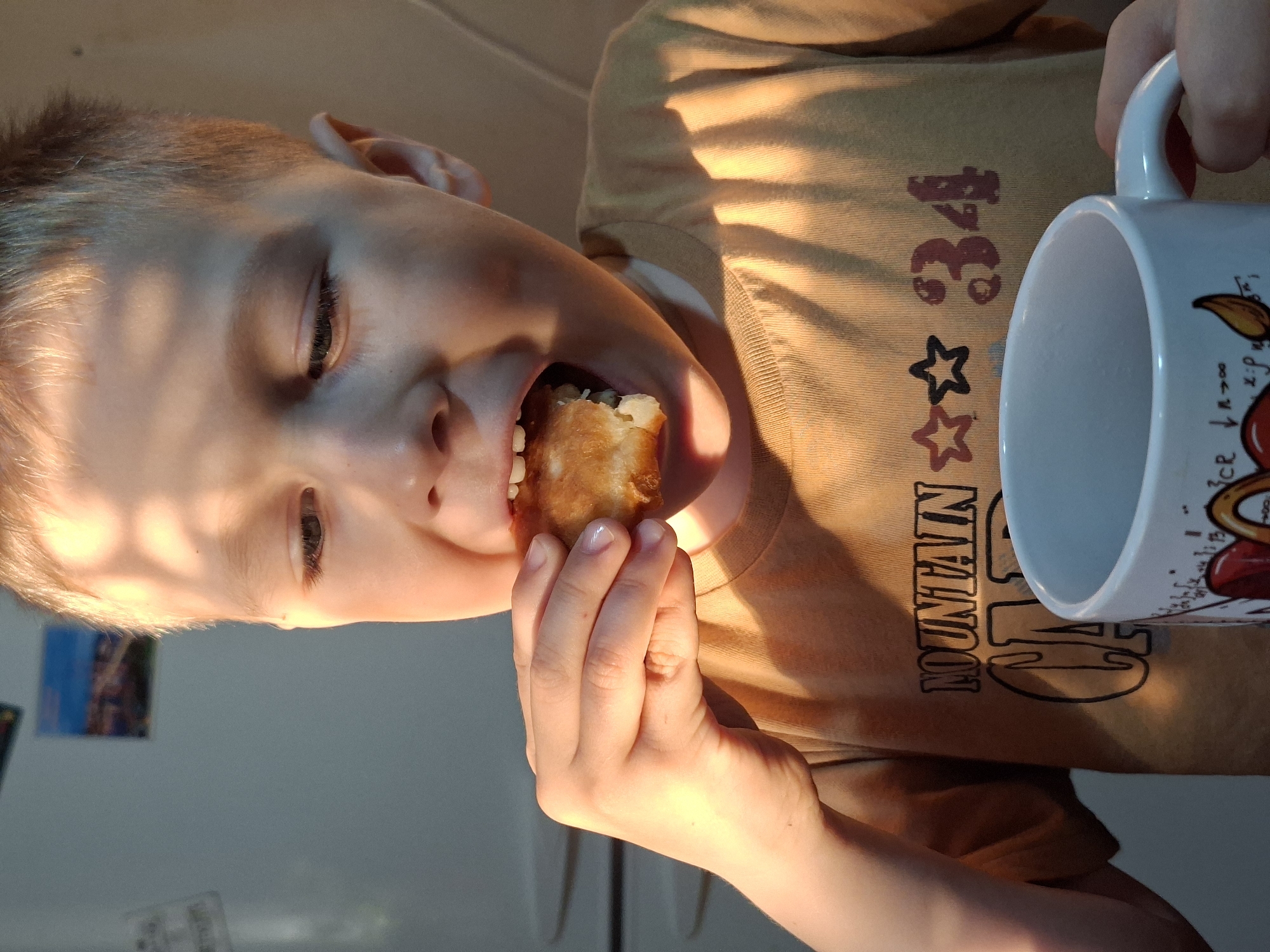 The image, rotated 90 degrees clockwise, captures a close-up of a young boy indoors, likely eating breakfast or a snack. He has light-colored hair and fair skin.
The central activity shows the boy biting into a golden-brown, pastry-like food item, which he holds in his right hand. His mouth is open, revealing his teeth and the food. With his left hand, he holds a white ceramic mug by its handle.
Notable details include the strong, warm, natural light illuminating the scene from the right, casting distinct linear shadows across his face, arm, and shirt. This suggests either early morning or late afternoon sunlight. The boy appears focused and content as he eats.
His clothing consists of a light brown or tan t-shirt. Visible text on the shirt includes the word "MOUNTAIN" printed vertically, along with stylized characters or numbers, possibly "34", and two stars (one outlined, one solid red). The white mug has a colorful design, part of which is visible, showing a red heart or fruit, swirly lines, and faint mathematical notation such as "n → ∞" and "x:p".
The background is a simple, light-colored wall. In the lower left, a small picture or magnet is visible on the wall, and a white appliance, possibly a refrigerator or cabinet, can be partially seen. The location is an indoor setting, likely a home kitchen or dining area.