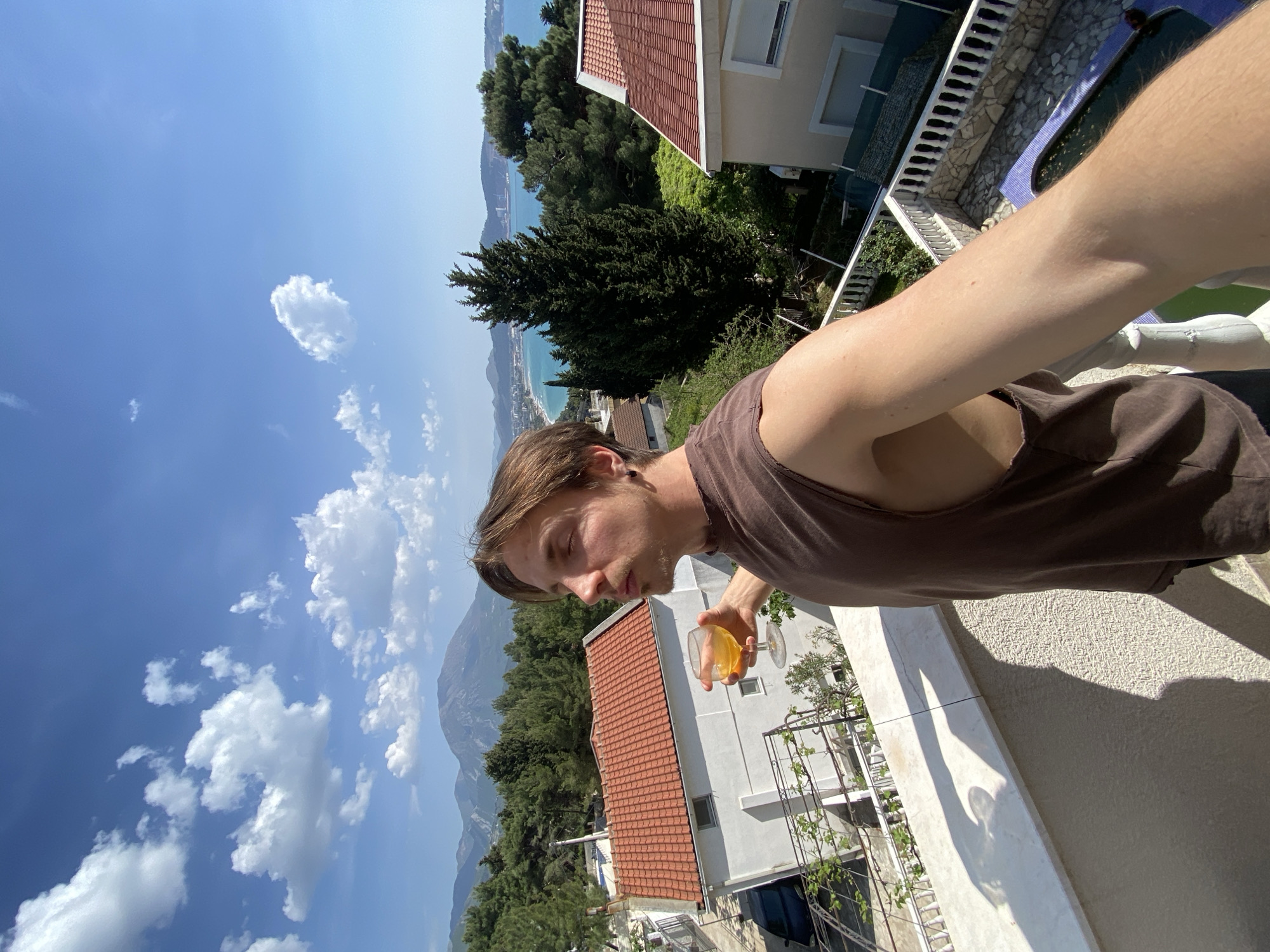 The image presents a young man on a sunny balcony overlooking the picturesque coastal town of Šušanj, Montenegro. In the foreground, a light-skinned man with medium-length brown hair and a light beard wears a dark brown sleeveless tank top. He holds a wine glass filled with a yellowish-orange liquid, looking down or to the side with a relaxed, contemplative expression, his eyes partially closed.
The background reveals a expansive view under a bright, clear blue sky dotted with scattered white cumulus clouds. Below the balcony, several white buildings with distinct red-tiled roofs are nestled among dense green trees, including tall conifers. Further down, parts of a blue swimming pool with white steps are visible. The vibrant blue sea stretches across the horizon, with a long, light-colored beach curving along the coastline. In the distance, majestic mountains rise, completing the scenic landscape. Strong shadows, such as the man's shadow cast on a light-colored, textured wall, indicate a midday or early afternoon setting. The scene conveys a sense of leisure and natural beauty. No visible text is present in the image.