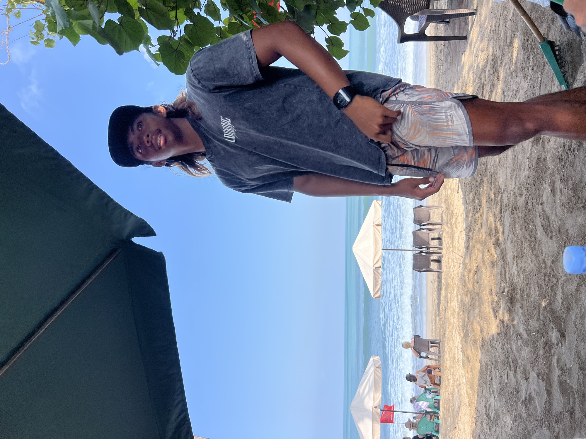 A young man wearing a dark t-shirt, patterned shorts, and a black baseball cap stands on a sandy beach in Kuta, Indonesia, looking up and to his right. He has long, dark hair and is wearing a smartwatch on his left wrist. The beach is lined with lounge chairs and umbrellas, with the ocean visible in the background. Several people are seated under umbrellas further down the beach, and a red flag is planted in the sand near one of the umbrellas. Lush green foliage is visible to the right of the man, and a rake is on the sand in the foreground. The sky is clear and blue.