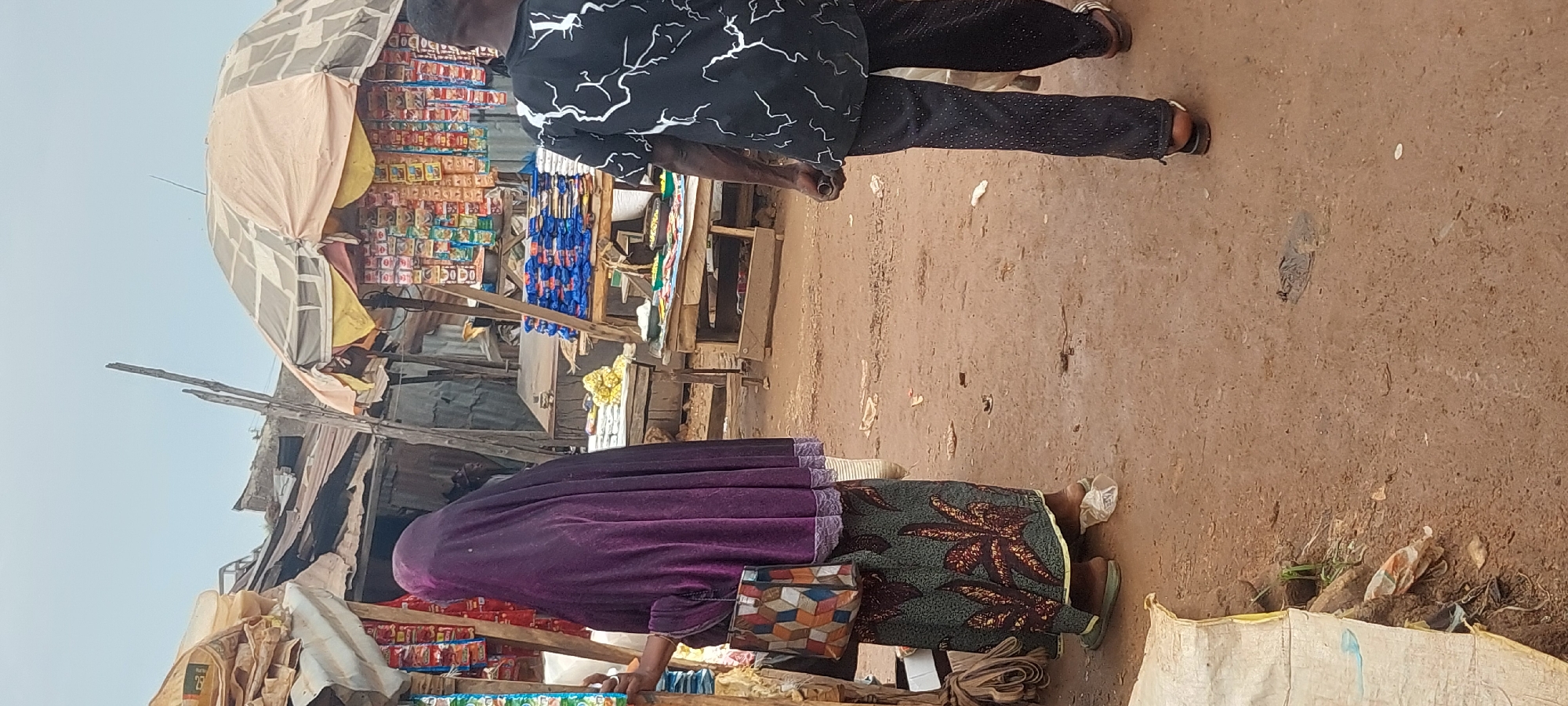 In a market setting with dirt ground and open-air stalls, two people are visible. On the left, a person wearing a purple headscarf and a patterned skirt stands facing away from the camera, holding a colorful bag. On the right, a person in a black shirt with white lightning bolt patterns and dark pants walks away from the camera. Various goods, including packaged snacks and produce, are displayed on wooden shelves under makeshift awnings. The environment appears somewhat unkempt, with some debris visible on the ground.