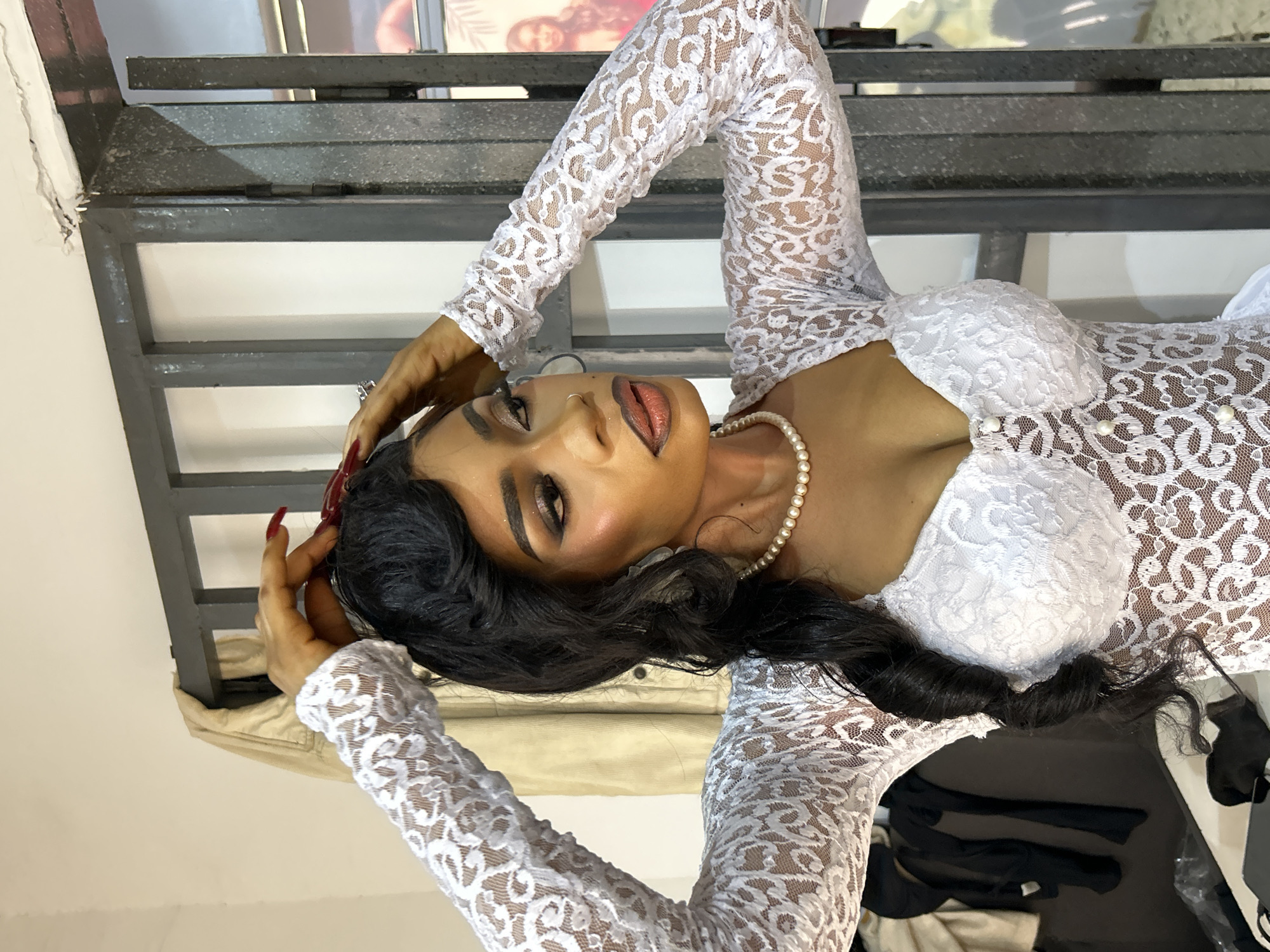 A woman is posing in Jos, Nigeria. She is wearing a white lace dress with long sleeves and a pearl necklace. Her hair is styled in dark, wavy curls. She has long red nails. Her hands are near her head. A metal structure with horizontal bars is behind her. There are various objects in the background including clothing.