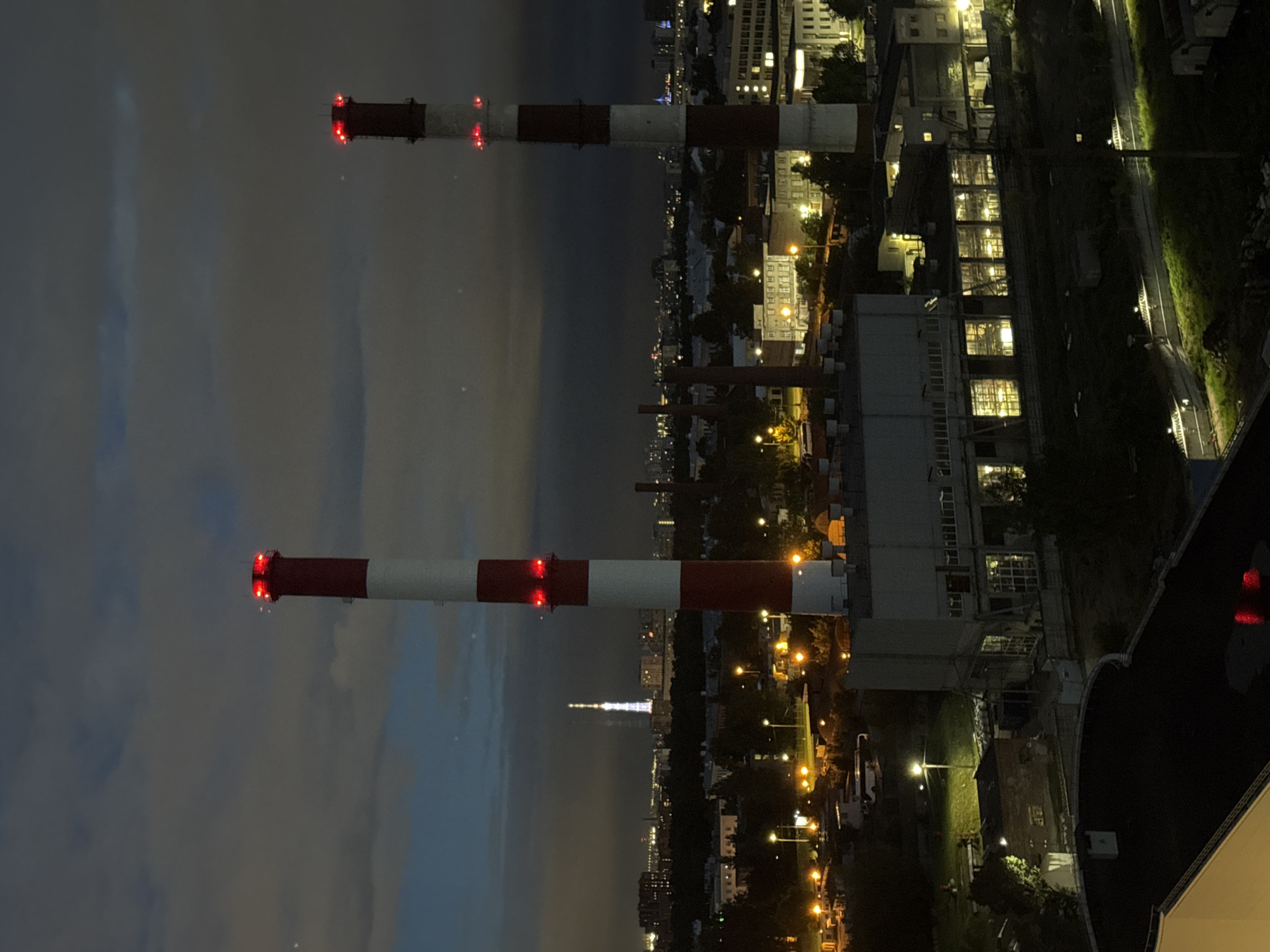 The photograph captures an aerial view of an industrial area at dusk, characterized by a dark, cloudy sky and artificial lighting. Two prominent smokestacks, painted in alternating bands of red and white, dominate the foreground. The taller smokestack on the left features multiple red lights at its summit and midway down, indicating operational status or hazard marking. The smokestack on the right also displays red lights near its top.
Below the smokestacks, a complex of industrial buildings is visible, with some structures illuminated by interior lights, suggesting activity within. The buildings are predominantly rectangular, with visible window panes and architectural details indicative of factories or processing plants. In the background, the cityscape stretches out, with numerous buildings and structures dimly lit against the darkening sky. A tall, illuminated tower, possibly a television or radio mast, stands out in the mid-distance, its structure outlined by a series of lights.
The ground level is sparsely detailed due to the aerial perspective and low light conditions, but hints of greenery, roads, and smaller structures can be observed. A bus is parked on a road to the left of the main industrial building. The overall atmosphere is one of urban industry operating into the night, with the red lights on the smokestacks serving as stark beacons against the twilight. The location is identified as Unknown, Russia.