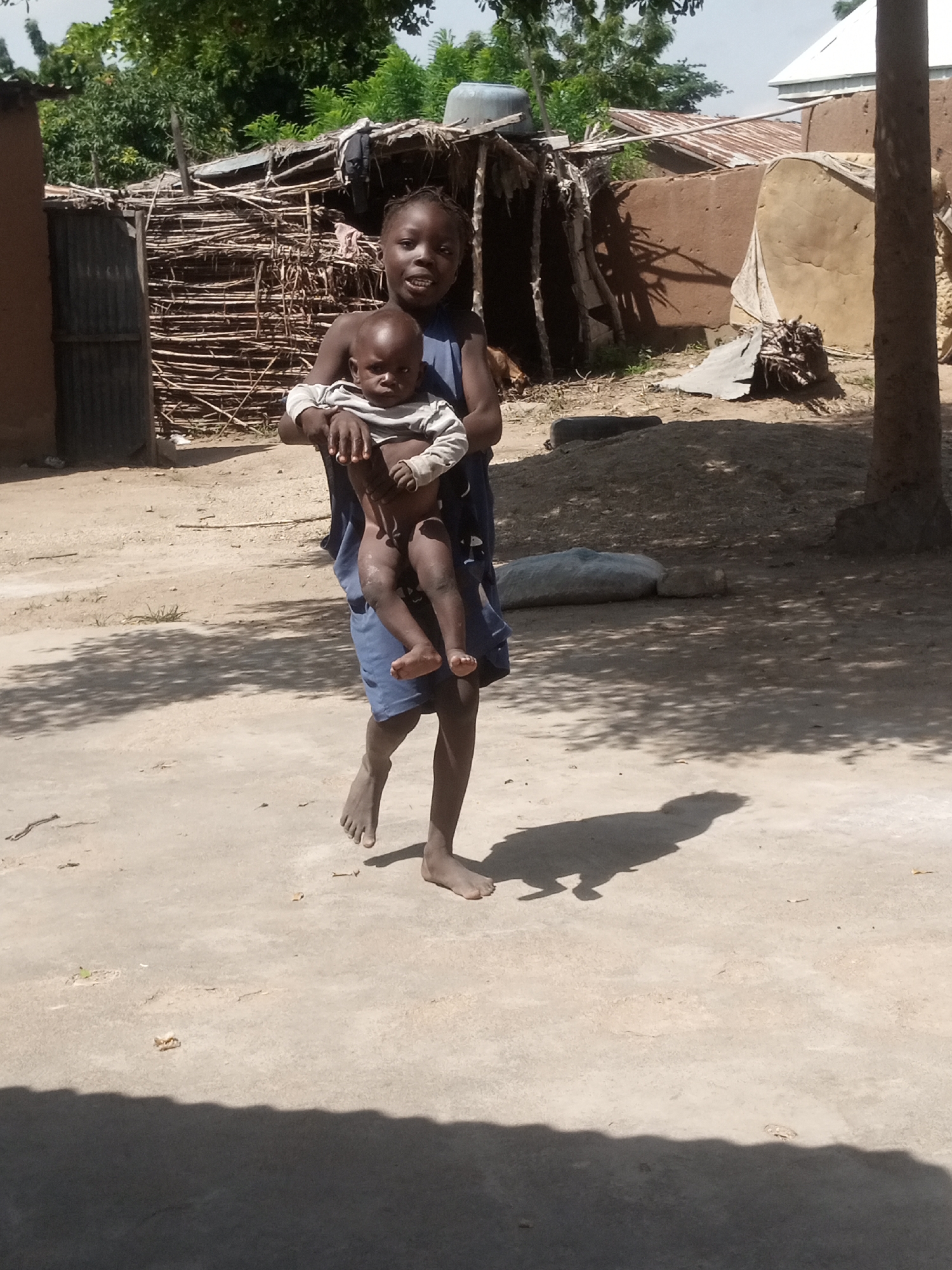 An outdoor, ground-level shot captures a young Black girl in a blue dress holding a dark-skinned baby. The girl is smiling and looking towards the camera, while the baby, wearing a grey and white striped shirt, gazes off to the side. They stand on a dirt path in a village setting. In the background, to the left, is a partially constructed mud-brick building with a corrugated metal roof. To the right, more mud-brick structures and thatched roofing are visible, along with a large tree trunk and some debris. The ground is a mix of sand and dirt, with scattered dry leaves and grass. Sunlight casts distinct shadows across the ground, suggesting a bright, sunny day. The overall impression is a snapshot of daily life in a rural Nigerian community.