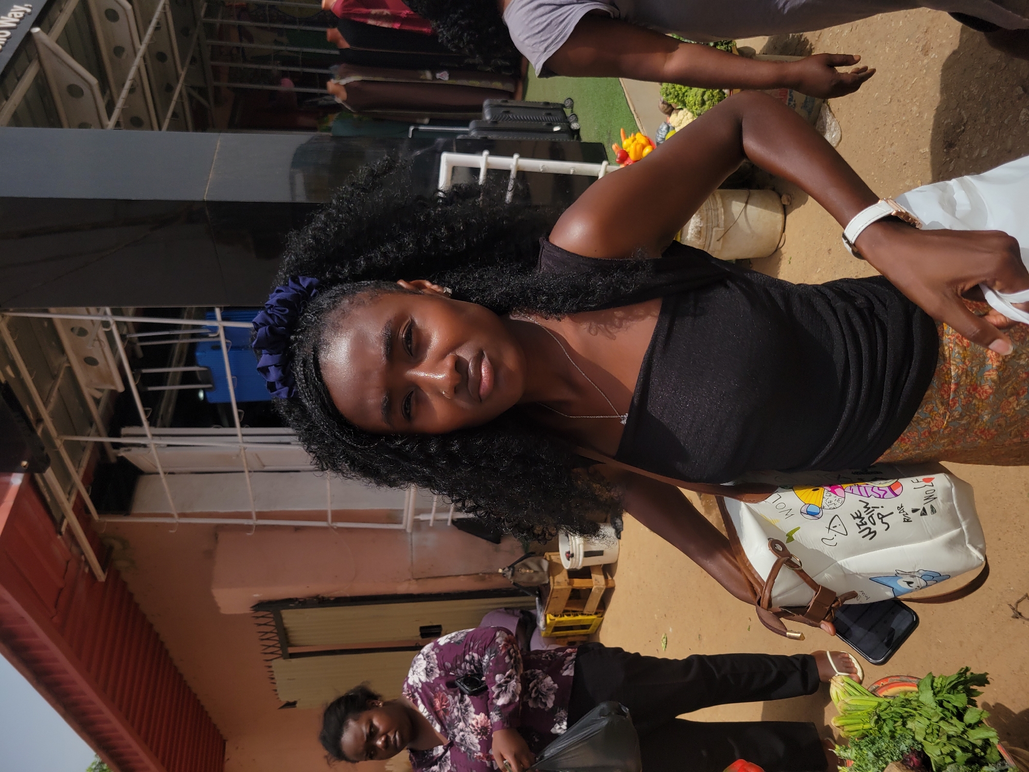 The image depicts a young Black woman, likely in her early to mid-twenties, standing in what appears to be an outdoor market or commercial area. The background features parts of buildings, possibly shops, indicating an urban setting. The woman is wearing a black top, with dark curly hair accented by a blue scrunchie, and a handbag over her shoulder. She is looking at the camera with a slight smile.
Another woman is visible in the lower part of the frame, seated with a bag and what looks like fresh produce, suggesting the market environment.
The image appears to be taken during daylight hours. Based on the metadata and contextual information, it's likely taken in Jos, Nigeria, given the architectural and market characteristics.